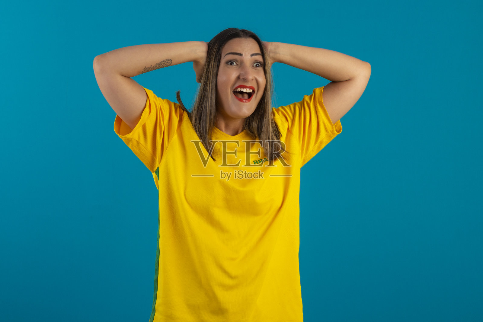 巴西女支持者,穿着印有巴西国旗的衬衫。巴西球迷在工作室的照片与蓝色背景和巴西的颜色。照片摄影图片