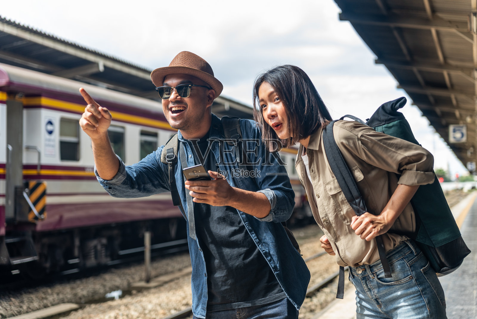快乐的亚洲帅哥和美女背包客戴着太阳镜,帽子和背包,当火车在火车站迎面而来时,他们指着和离开。照片摄影图片