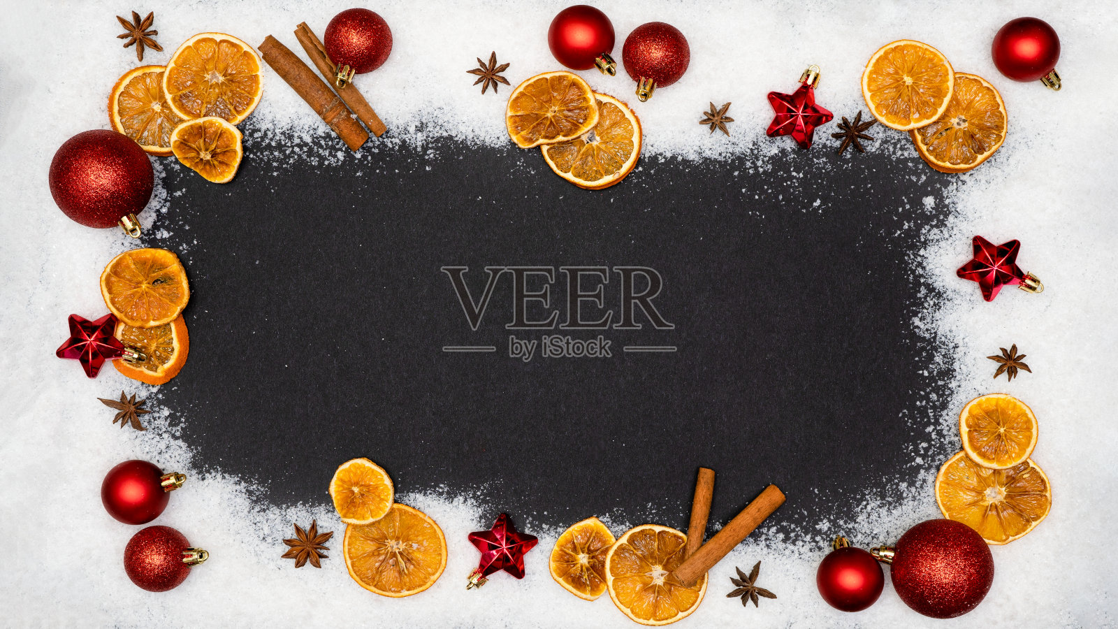 节日圣诞降临庆祝节日背景横幅贺卡-边界框架制成的圣诞装饰品,红色的小饰物,球,星星,干橙片,肉桂棒和雪上黑色的桌子纹理,顶视图照片摄影图片