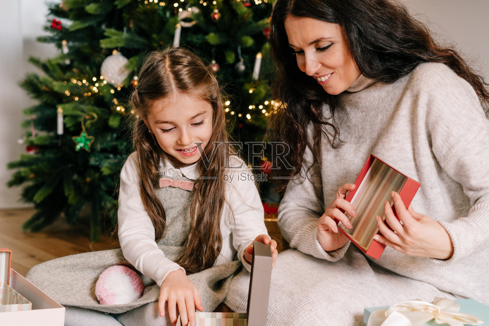 兴奋的女儿和母亲坐在家里装饰漂亮的圣诞树旁,享受打开他们的礼物。家庭度假。节日的圣诞气氛。有选择性的重点。照片摄影图片