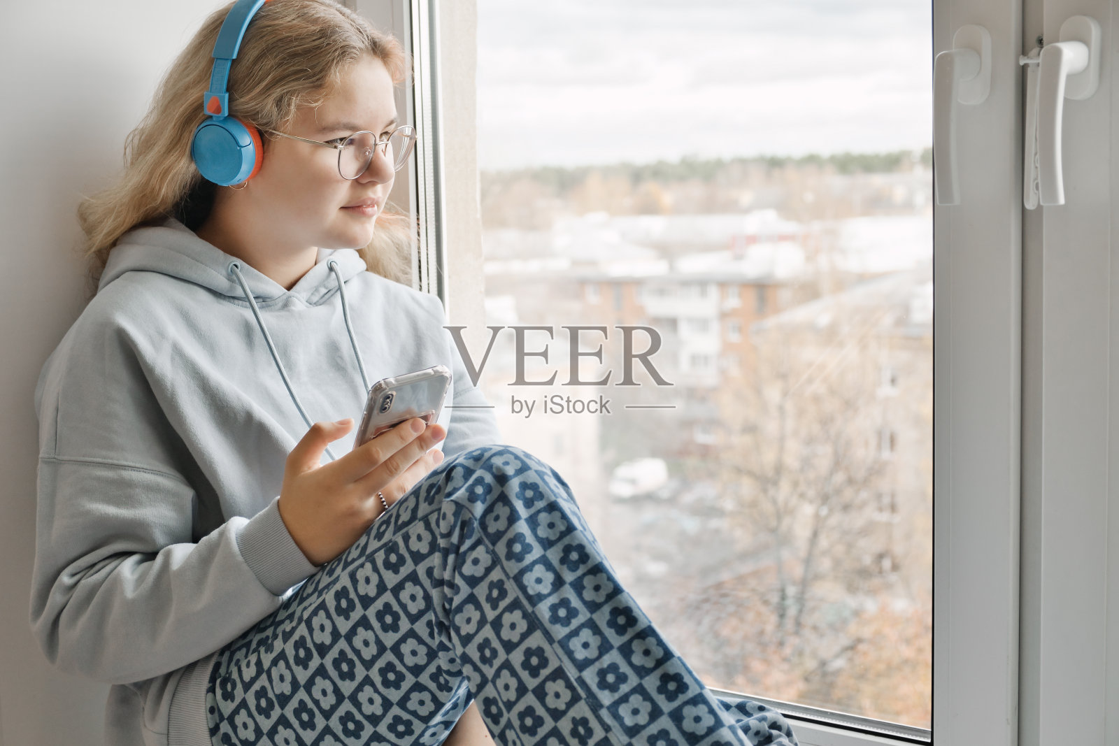 美丽的年轻女孩戴着眼镜,手里拿着智能手机,坐在窗台上,戴着耳机听音乐。照片摄影图片