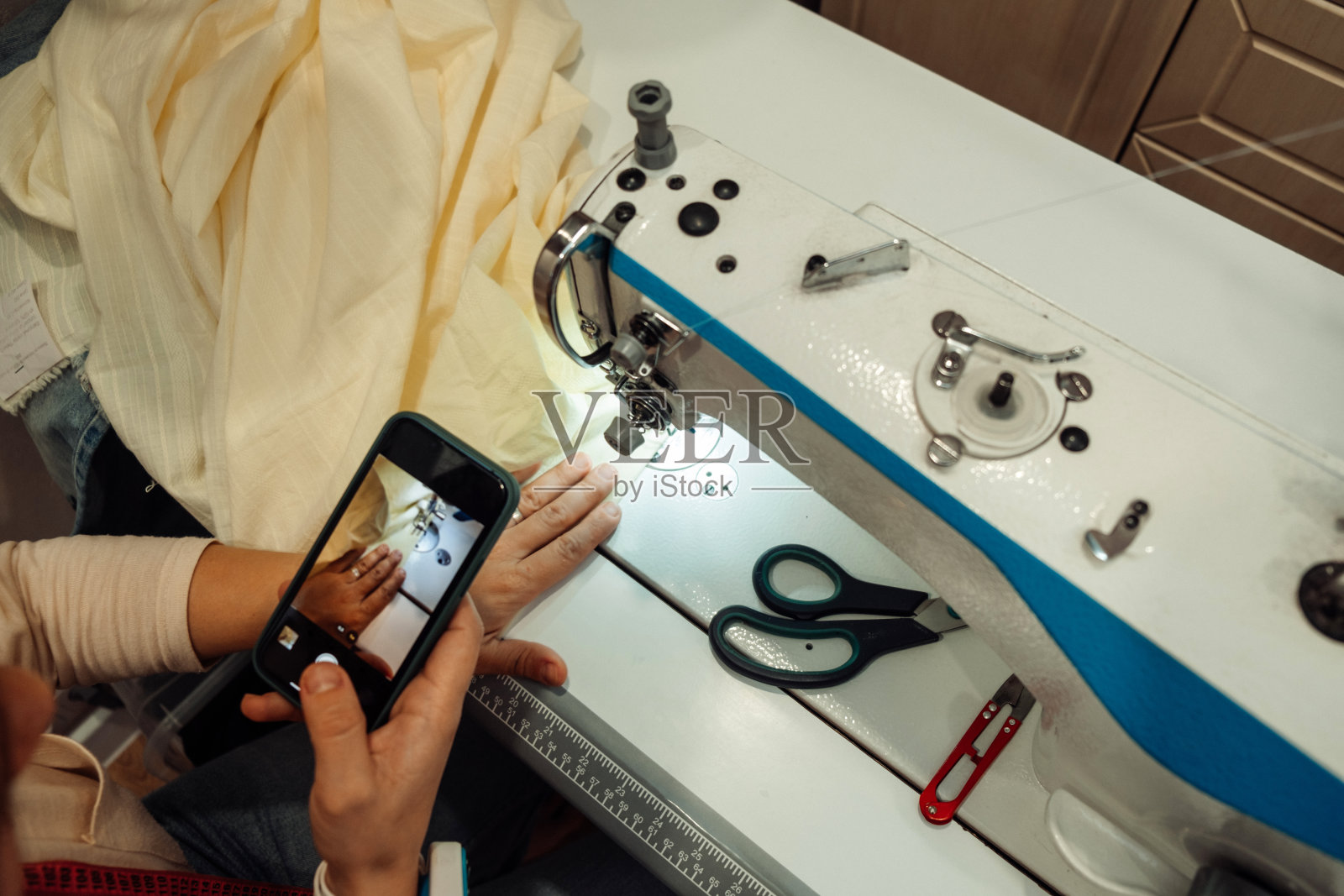 一名白人妇女在家庭作坊的缝纫机上缝制衣服,用智能手机用特写镜头录下了直播。小生意。博客的概念。照片摄影图片