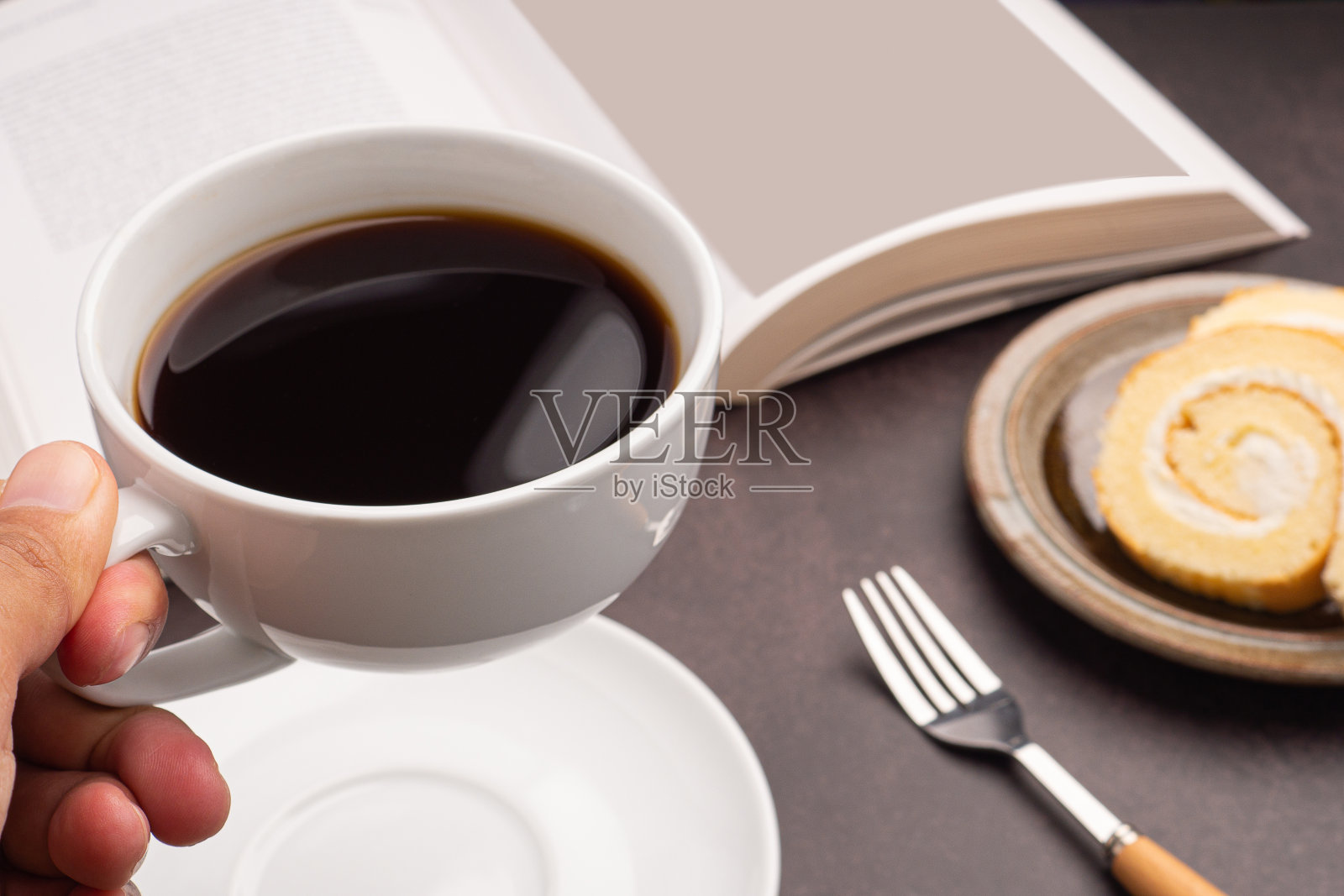 手里拿着一个白色的咖啡杯和一本书,盘上卷着蛋糕照片摄影图片