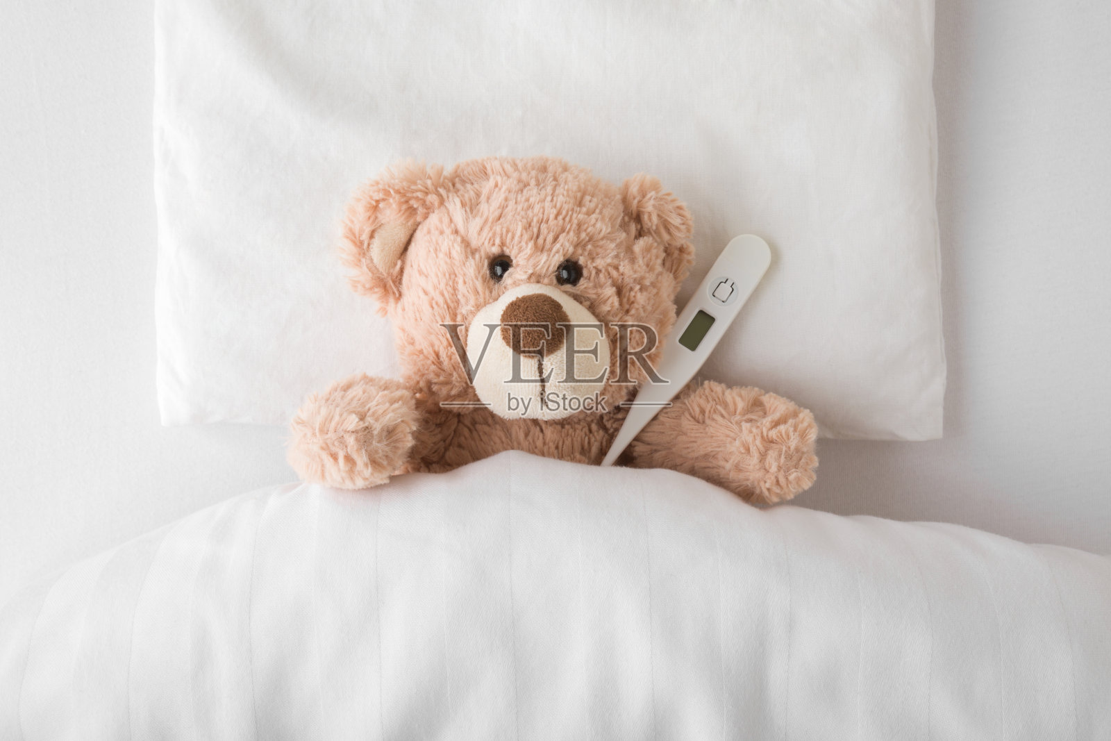 在白色的床上,带着数字温度计的泰迪熊躺在枕头和床单下。在感冒和流感病毒时期测量体温。儿童保健概念。特写镜头。自顶向下视图。照片摄影图片