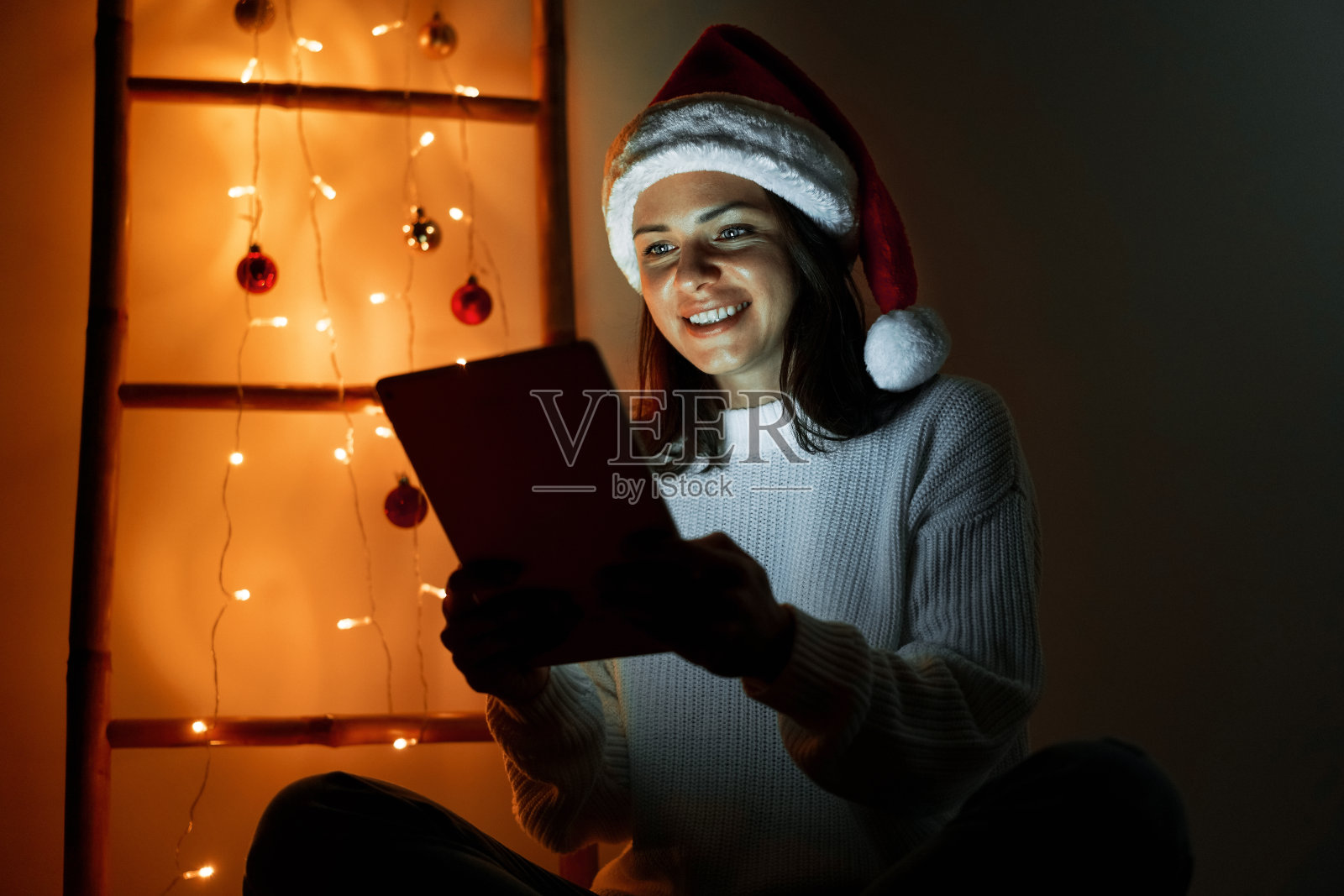 戴着圣诞帽的妇女在家中使用数字平板电脑,在圣诞夜平板电脑照亮了妇女的脸照片摄影图片