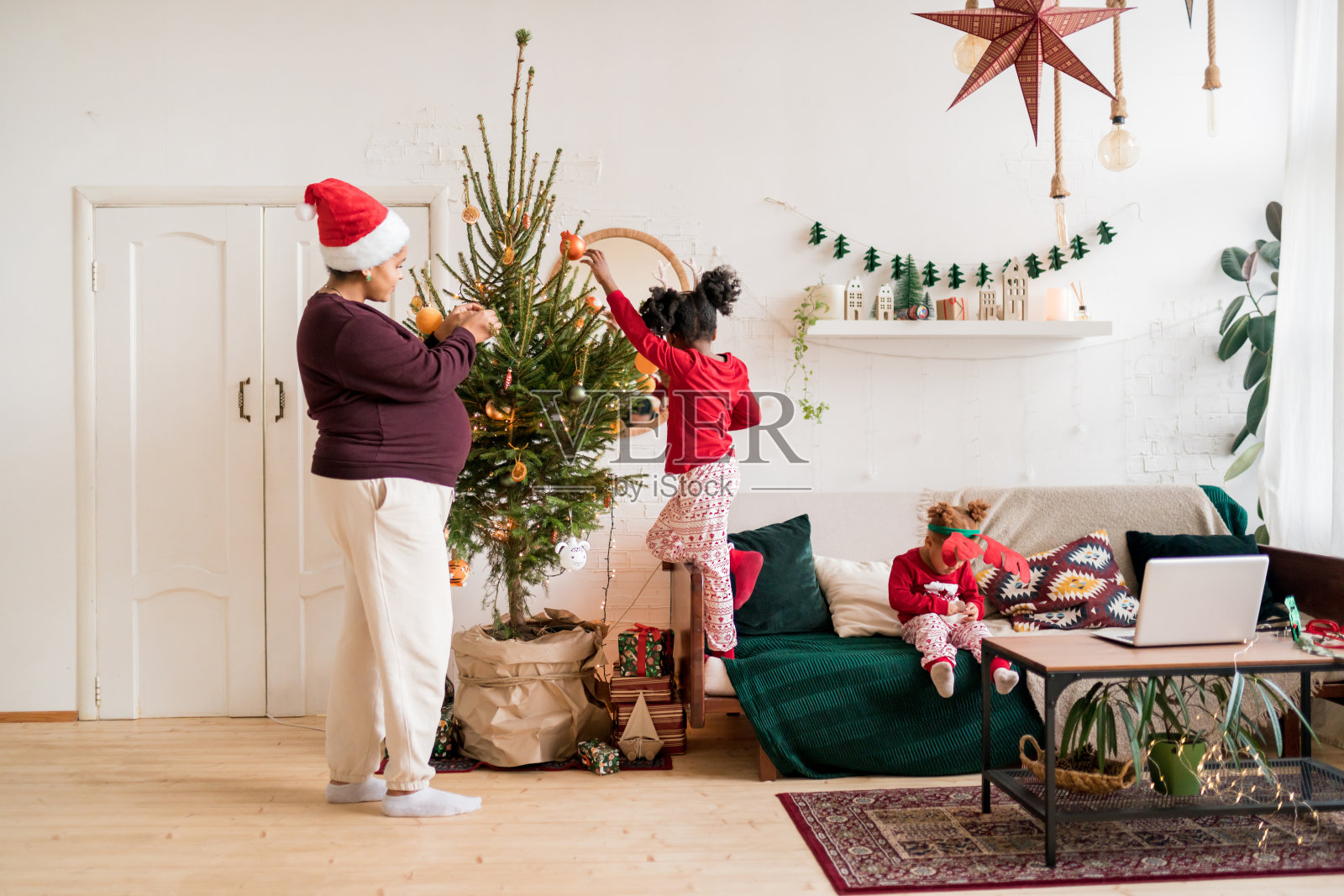 身着圣诞睡衣的非裔美国女孩在家中装饰着怀孕母亲的圣诞树。与孩子在家共度圣诞时光的概念照片摄影图片