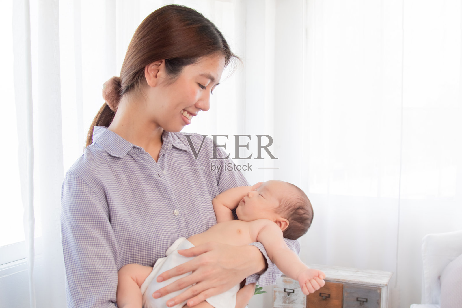 美丽的亚洲妈妈抱着刚出生的婴儿,婴儿在她的怀里睡觉,快乐的妈妈站在家里,用爱和保护哄可爱的婴儿。拥抱表示爱和关心,母亲的概念。照片摄影图片
