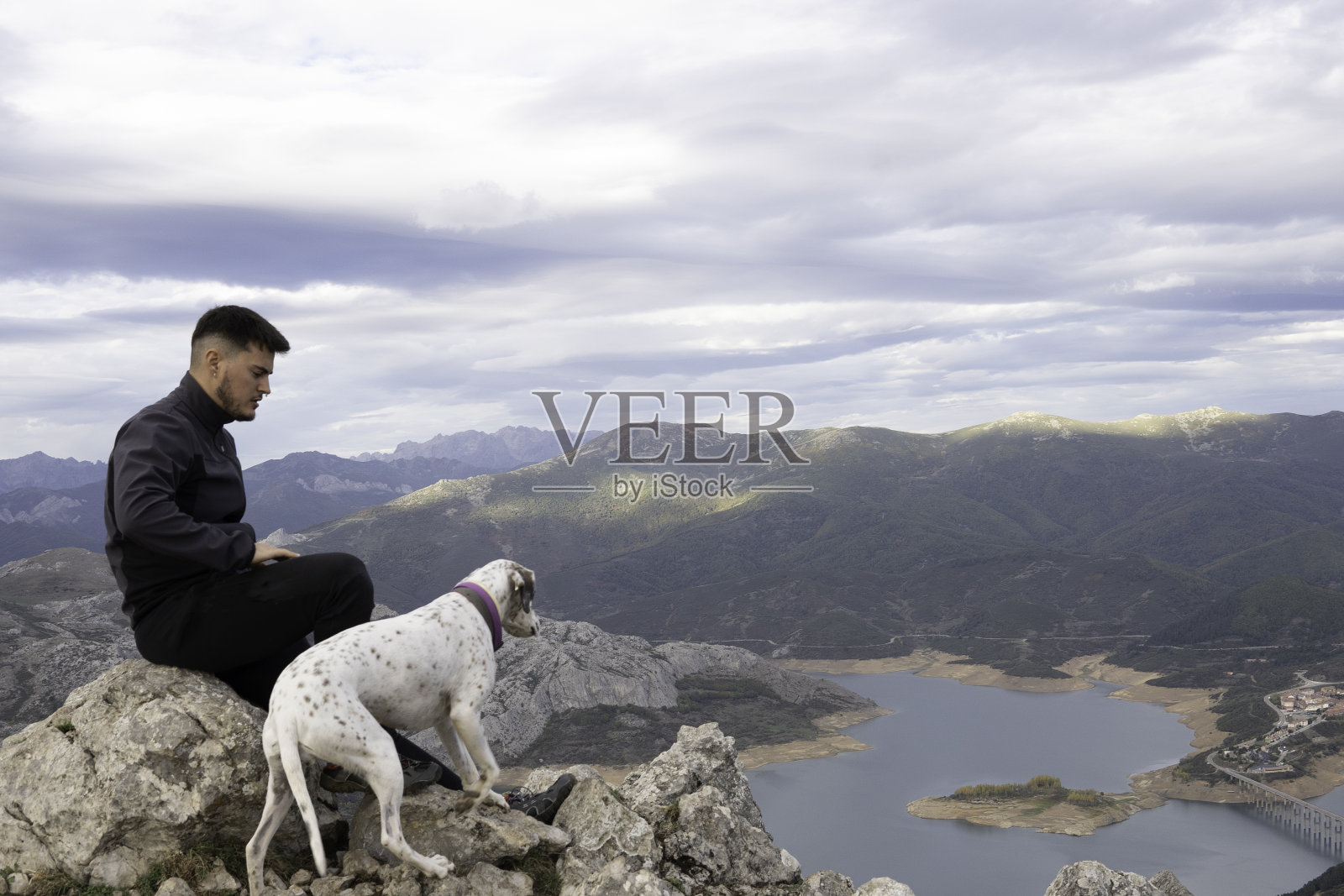 在山顶上,一个穿着深色山装的年轻登山者在岩石上休息,旁边是他的狗。在背景中我们可以看到一个湖和附近的山脉。徒步旅行和登山的壁纸。照片摄影图片