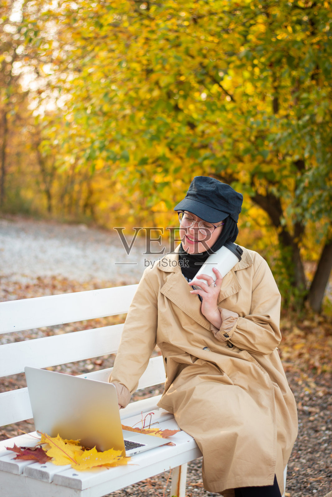 在公园里,一名戴着头巾、穿着米色风衣的穆斯林妇女坐在长凳上,手里拿着一杯咖啡和一台笔记本电脑。穆斯林女商人。学生。视力用眼镜。照片摄影图片