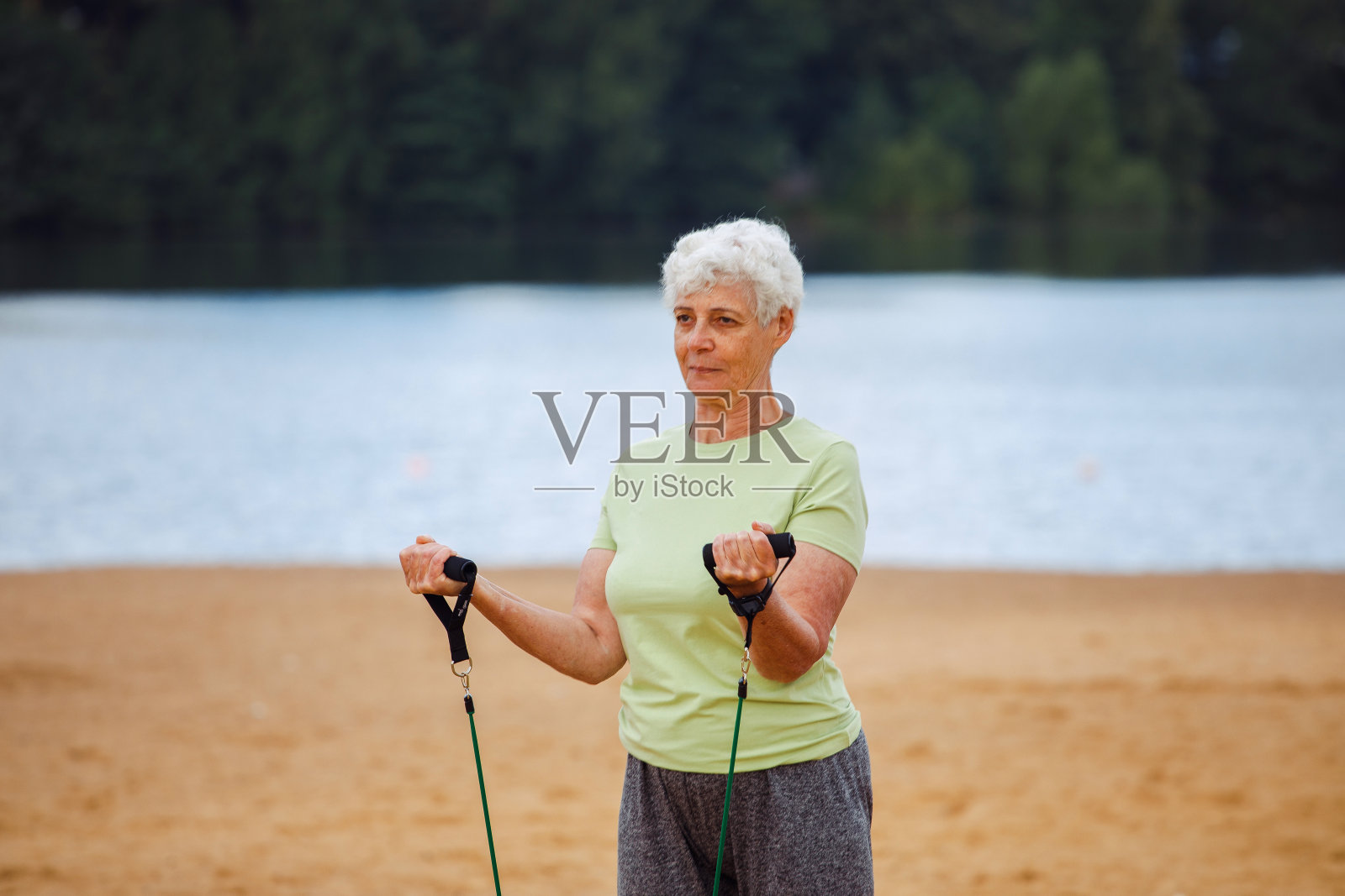 早晨,穿着运动服的老妇人在沙滩上用橡皮筋做运动。健康的生活方式,积极的退休生活和运动时间。照片摄影图片