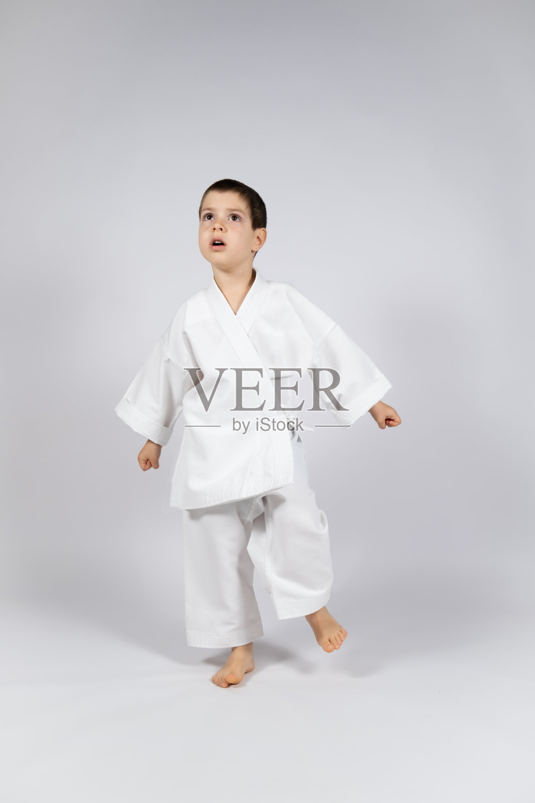 一个穿着和服的小男孩在练习空手道,抬头看着白色背景。照片摄影图片
