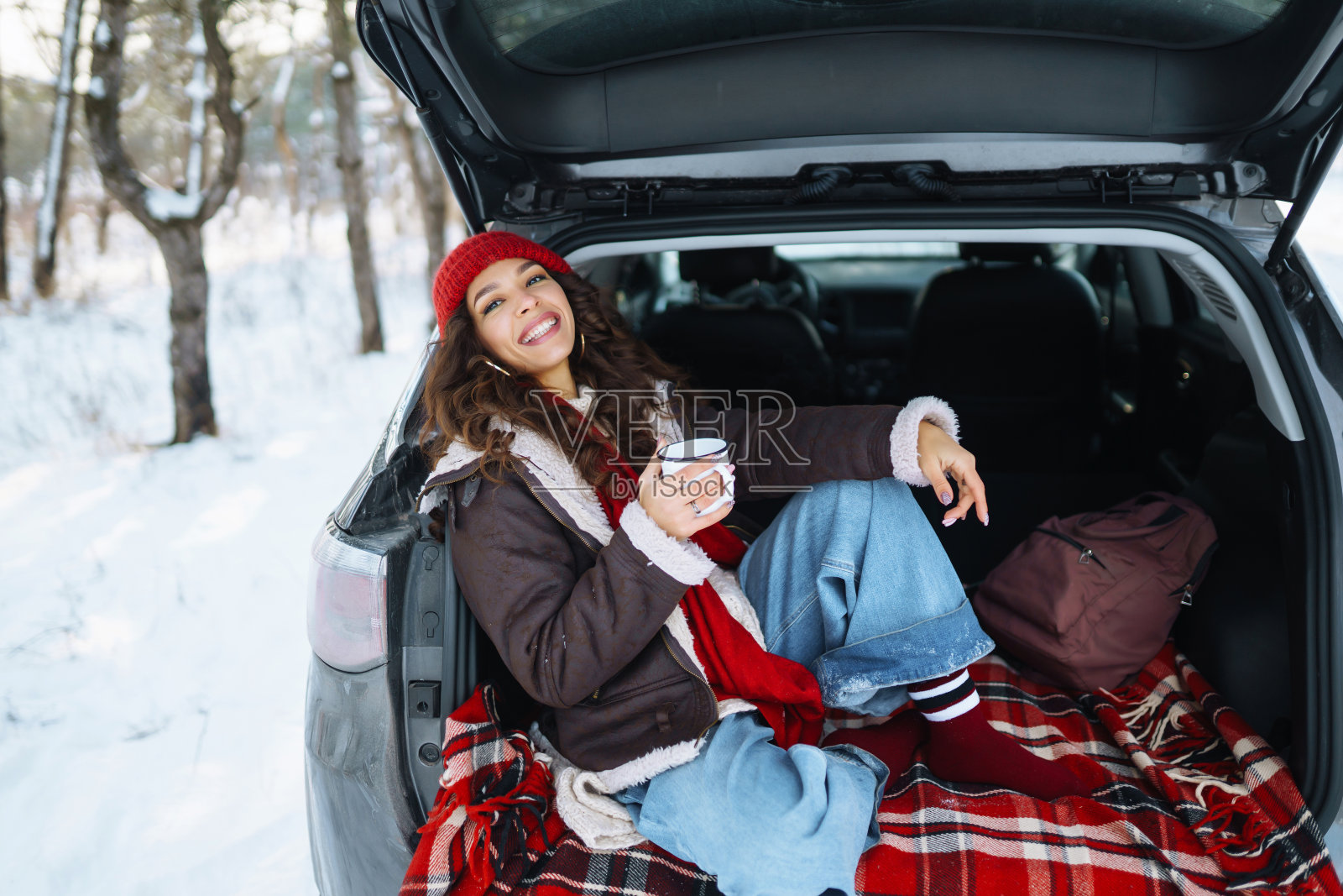 在冬天的森林里,快乐的女人拿着保温杯坐在汽车后备箱里喝茶。照片摄影图片