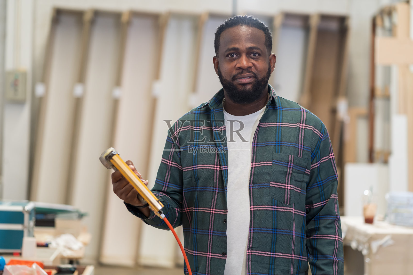 非裔美国木匠持有气动钉枪在木材车间。木工男工人肖像在安全制服和设备工具与气动钉枪工作在木材工厂照片摄影图片