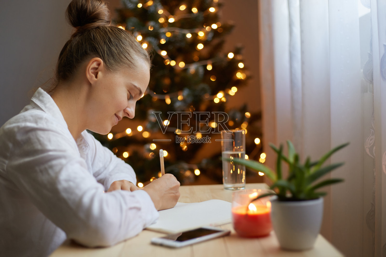 穿着白衬衫的微笑快乐的女人坐在桌子旁,写着圣诞节遗愿清单或她需要做的寒假准备工作,在有圣诞树的房间里写在笔记本上。照片摄影图片
