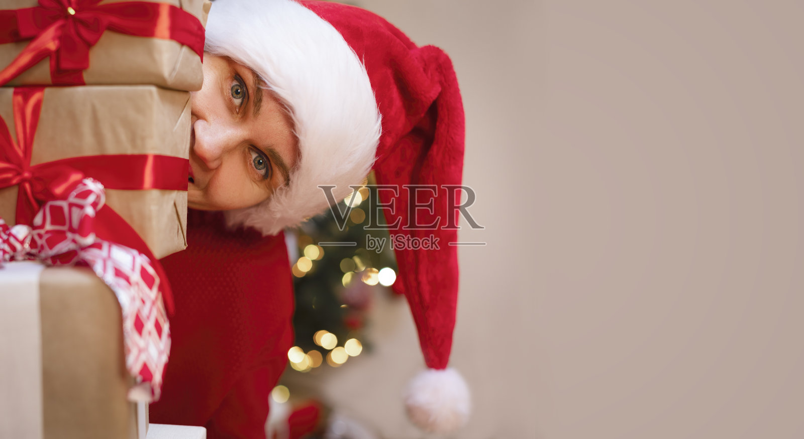 一个戴红帽子的女孩从圣诞礼物堆里探出头来,微笑着。照片摄影图片