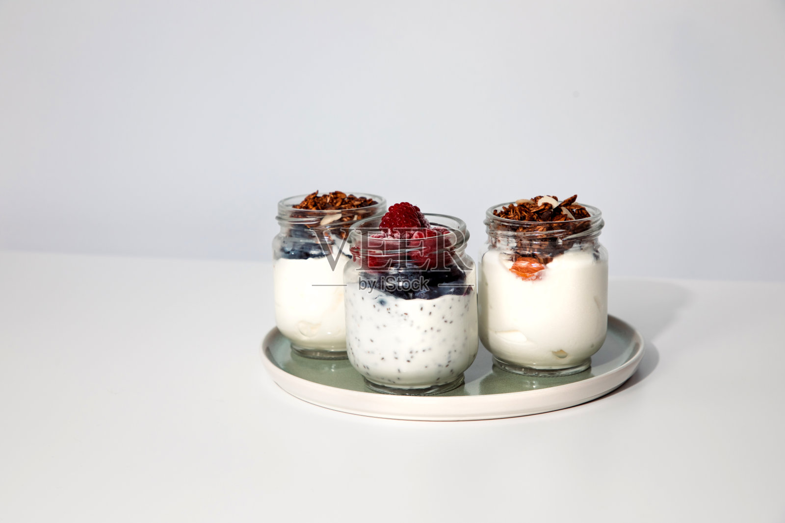酸奶装在玻璃罐里,蓝莓、覆盆子、奇亚籽和格兰诺拉麦片放在白色背景的绿色盘子里照片摄影图片