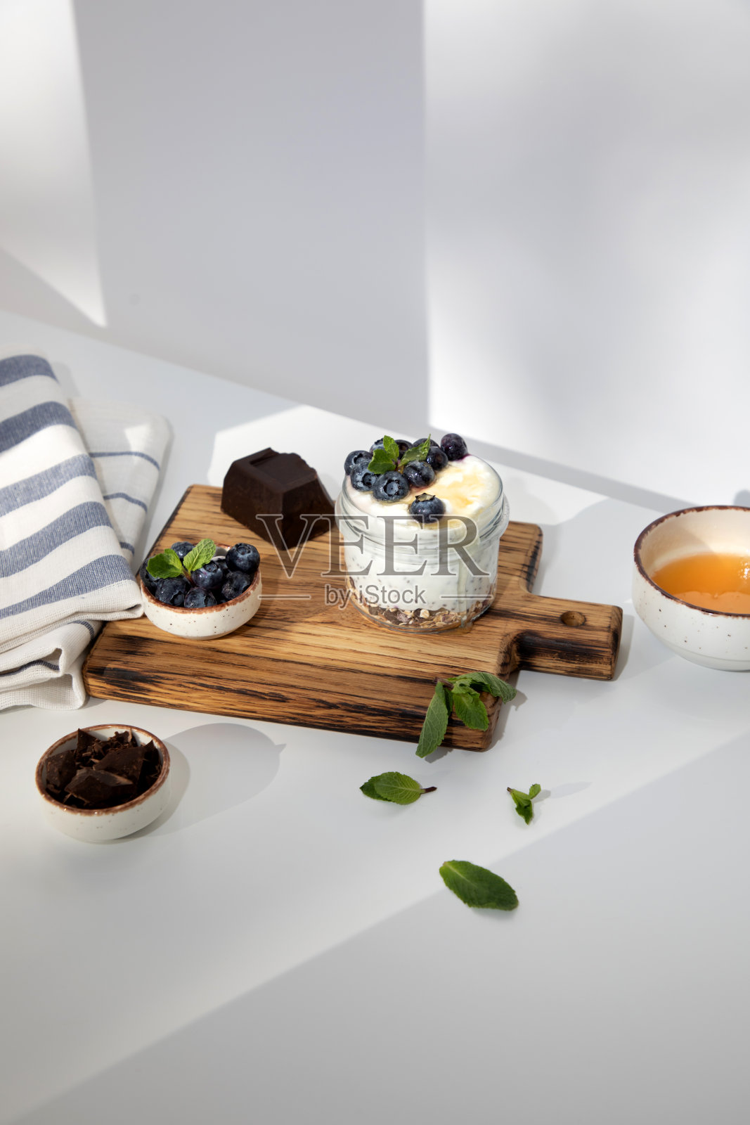酸奶装在玻璃罐里,配上格兰诺拉麦片、巧克力、蓝莓和蜂蜜,放在木板上,白色背景上放上薄荷叶照片摄影图片