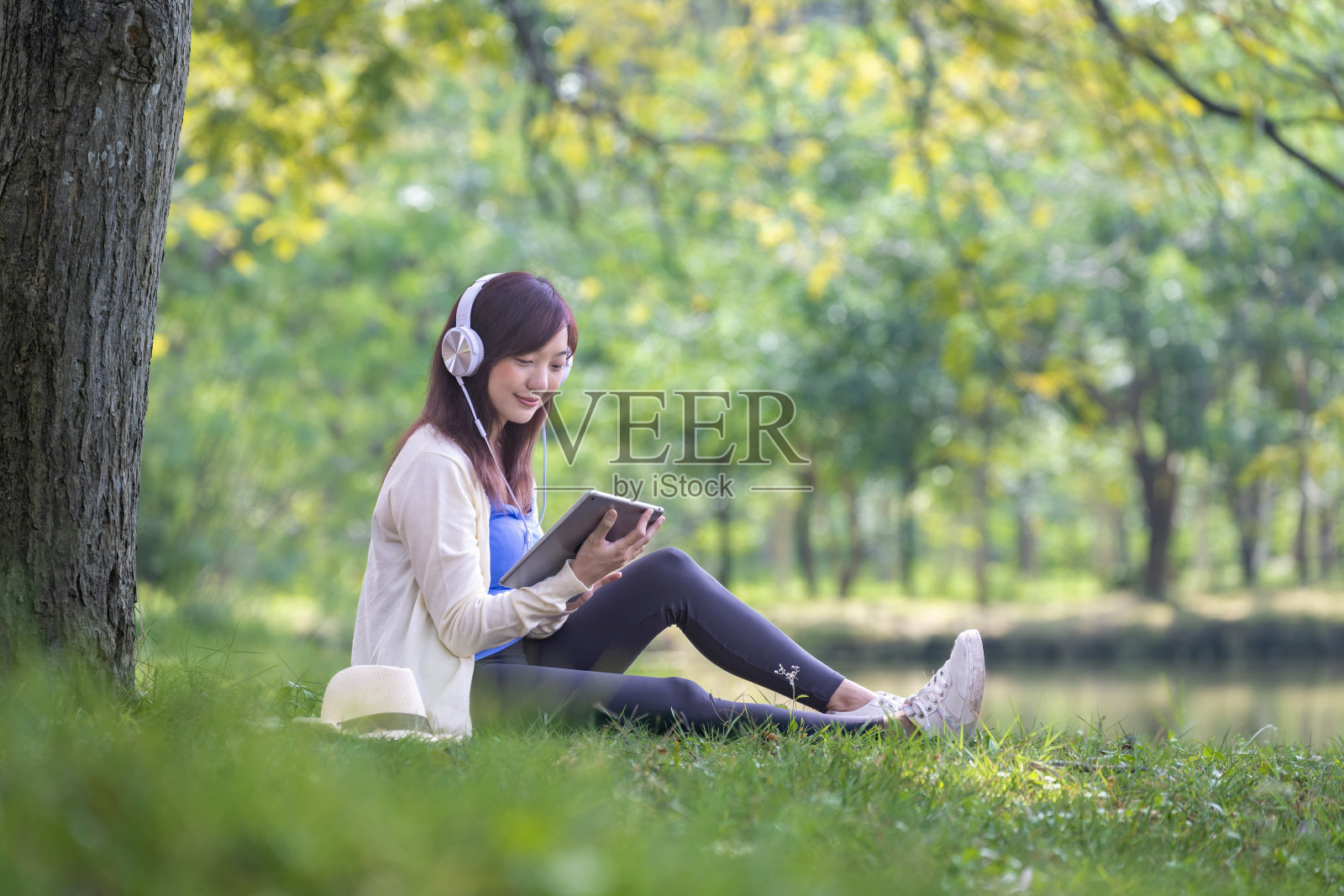 图为,一名年轻的亚洲大学生正坐在大学校园自然公园湖边的树下,聆听有声读物,在大自然中学习和放松照片摄影图片
