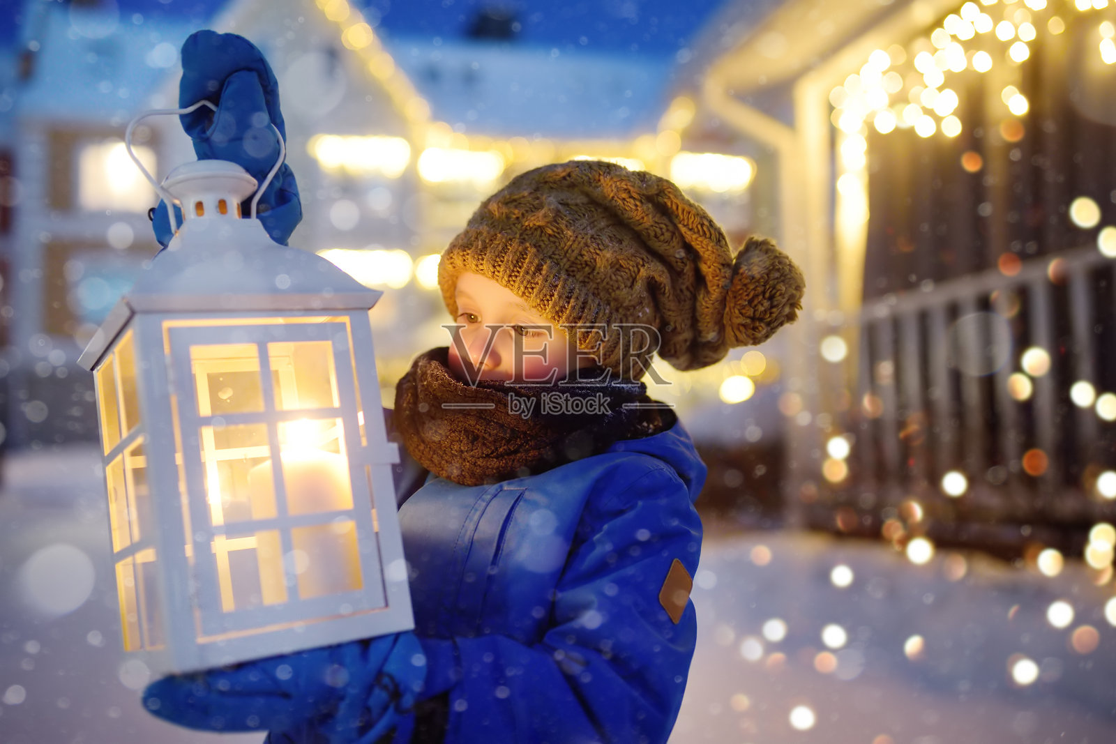 在圣诞夜的大雪中,一个小孩在小镇的街道上等待圣诞老人。男孩拿着点着蜡烛的大灯笼。婴儿希望魔法和礼物。照片摄影图片