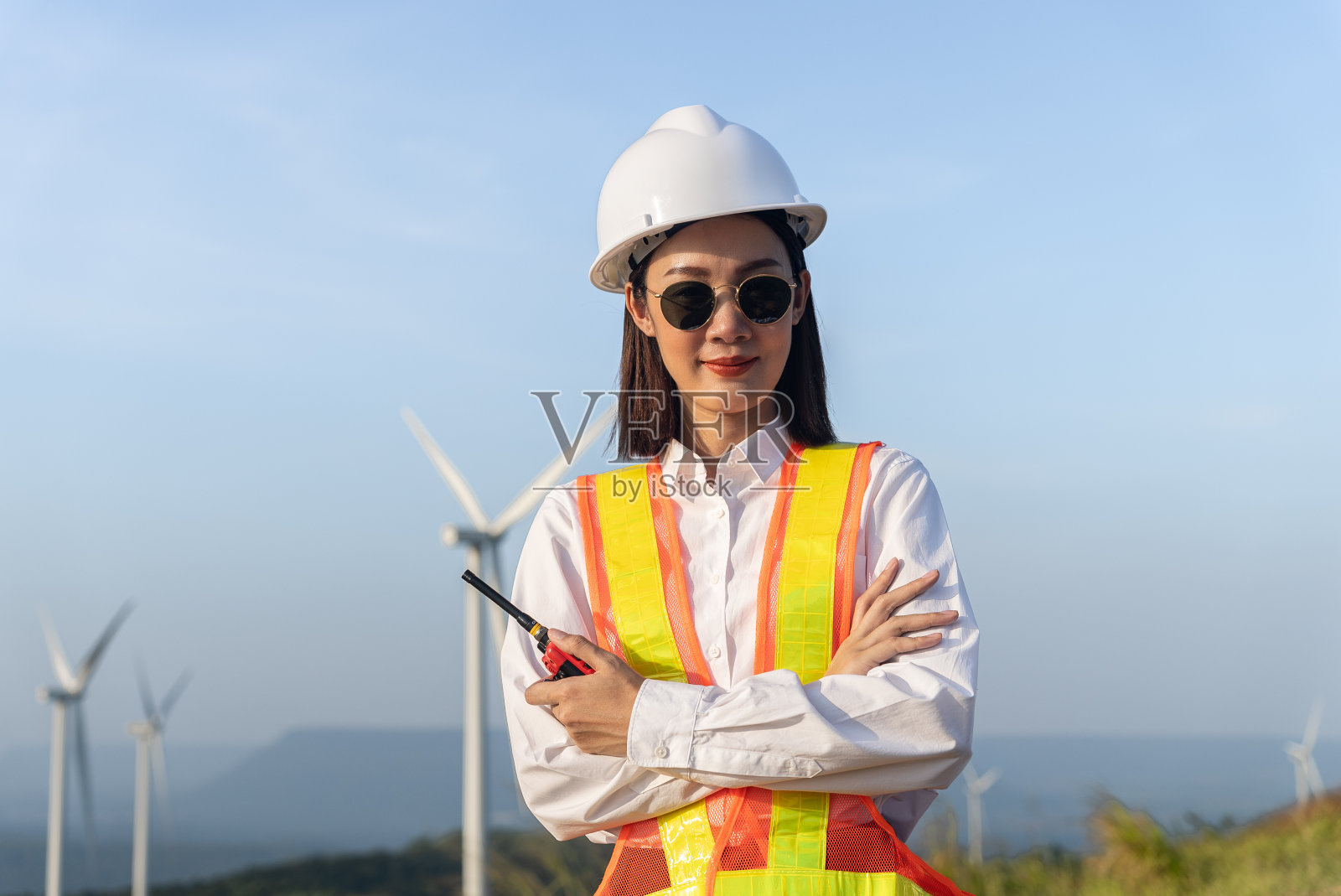 美丽的亚洲女工程师戴安全帽,太阳镜,反光背心和折叠的手臂和手持对讲机与风力涡轮机螺旋桨的背景肖像。环境友好。照片摄影图片