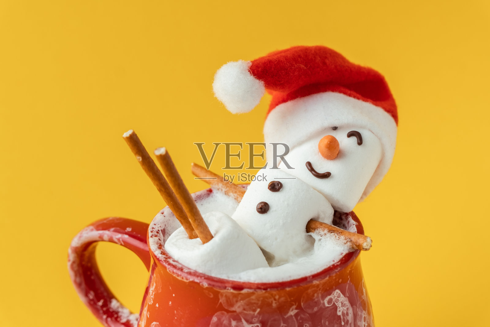 棉花糖雪人在一个充满可可和牛奶泡沫的红色陶瓷杯热浴缸。圣诞假期黄色背景。冬季的概念。热巧克力加棉花糖和节日装饰。照片摄影图片