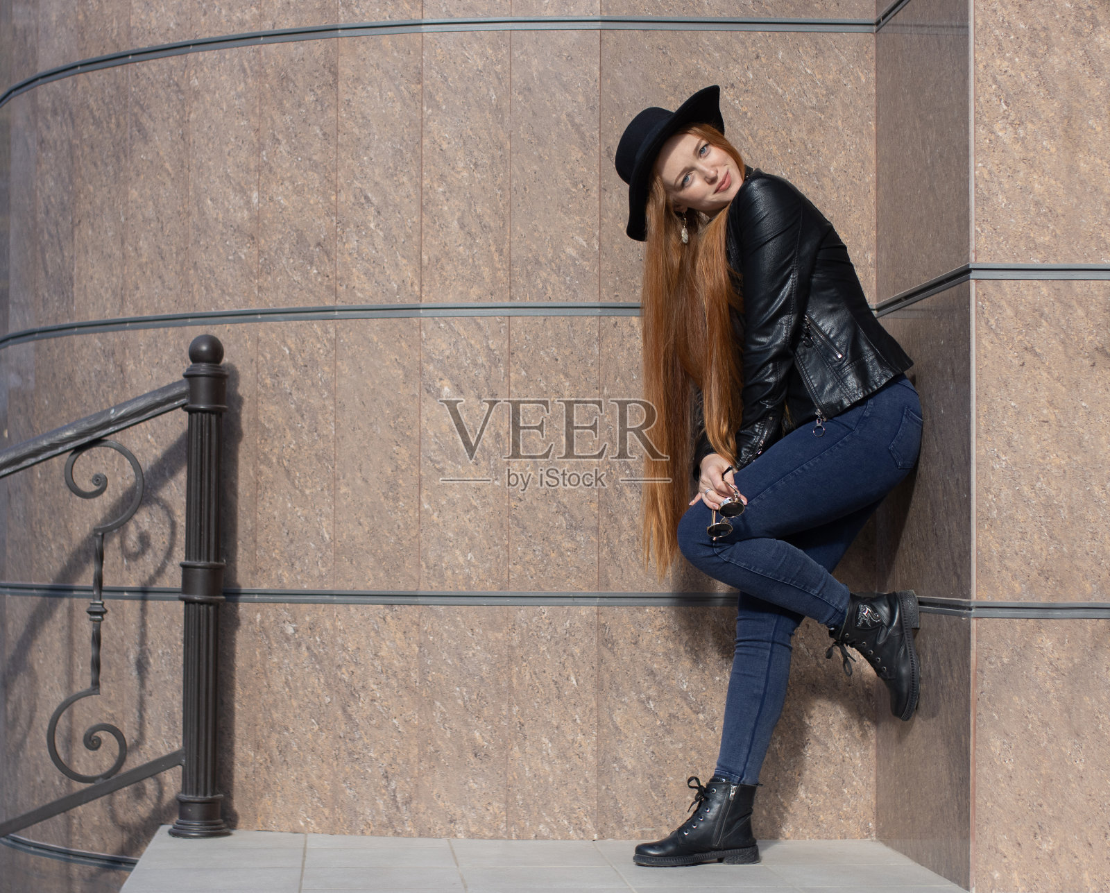 一个漂亮的长发女孩,戴着帽子,穿着皮夹克,在街上靠在一幢建筑的花岗岩墙上摆姿势。照片摄影图片