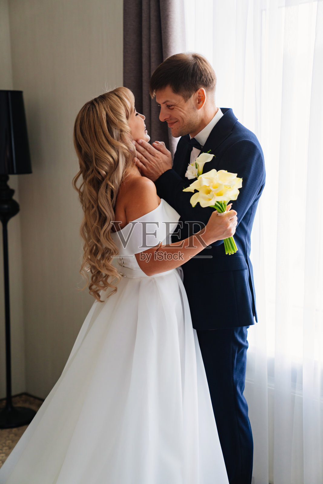 温柔相爱的金发新娘和新郎站在窗边。照片摄影图片