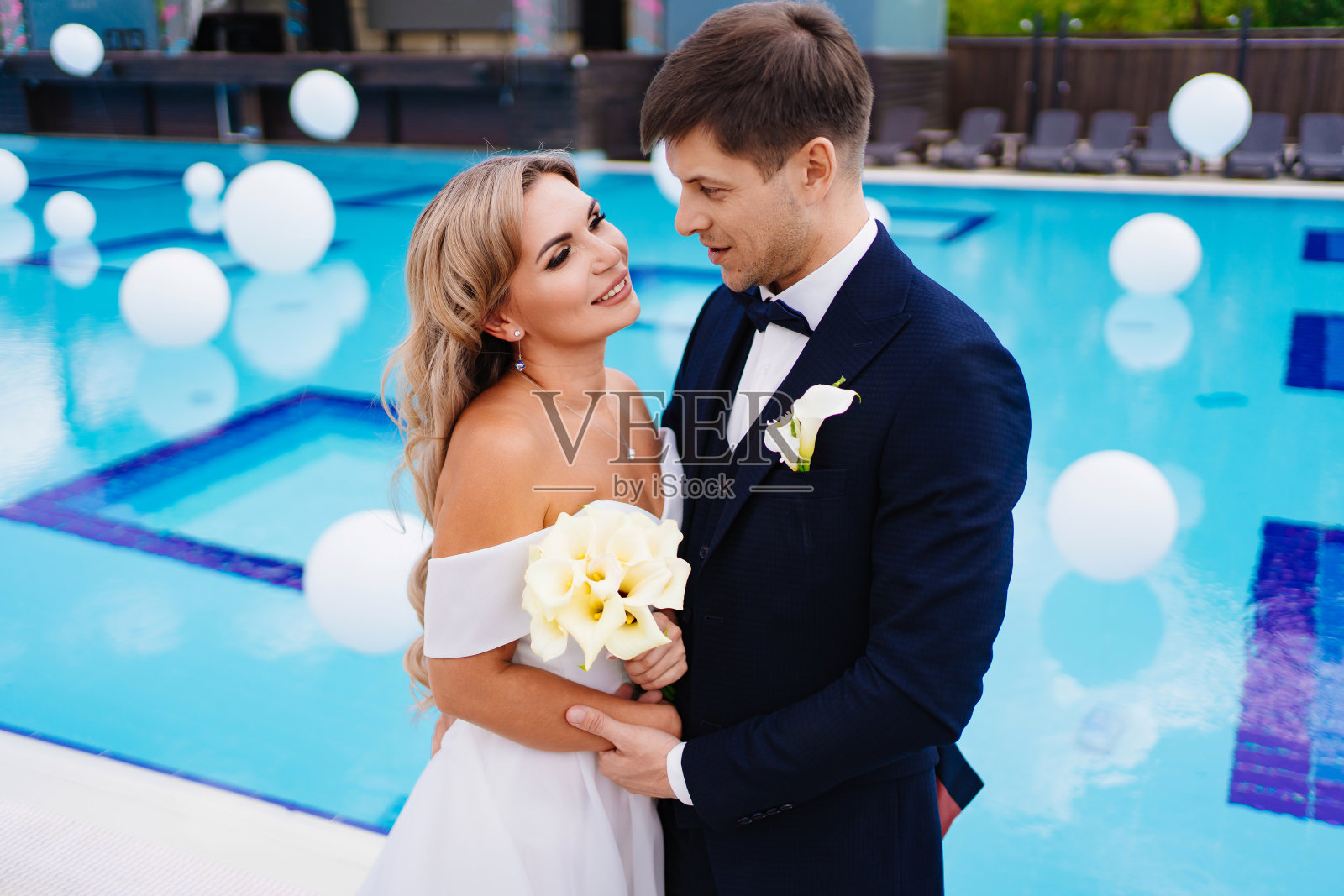 新娘和新郎穿着经典的衣服,站在装饰着白色气球的泳池边。照片摄影图片