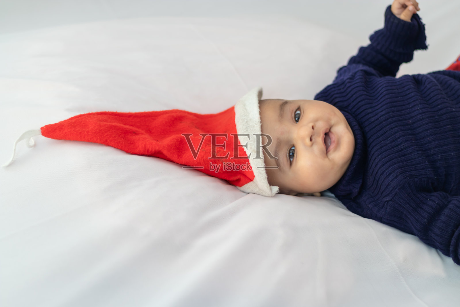 婴儿男孩戴着圣诞红帽躺在室内可爱的面部表情与白色背景照片摄影图片