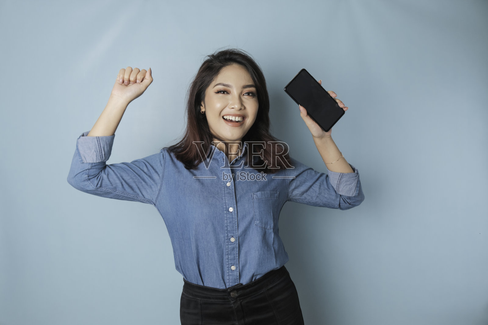 一名年轻的亚洲女性穿着蓝色衬衫,手拿被蓝色背景隔开的智能手机,表情快乐而成功照片摄影图片