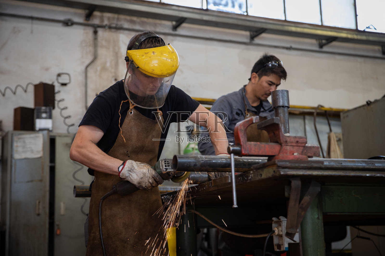 一名工人在工厂里用磨光机打磨一块金属,他的同事在旁边工作。照片摄影图片