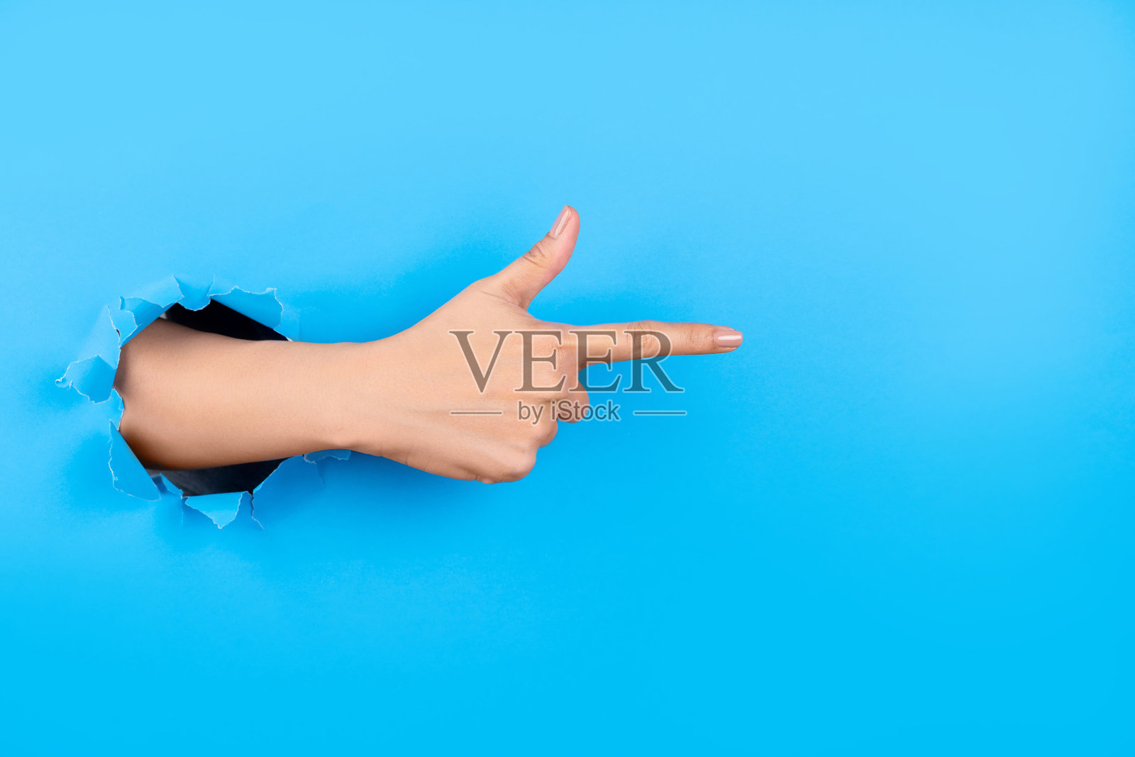 人手穿过纸墙,用食指在蓝色背景上指向虚拟物体或文本,为文本留出空间。照片摄影图片