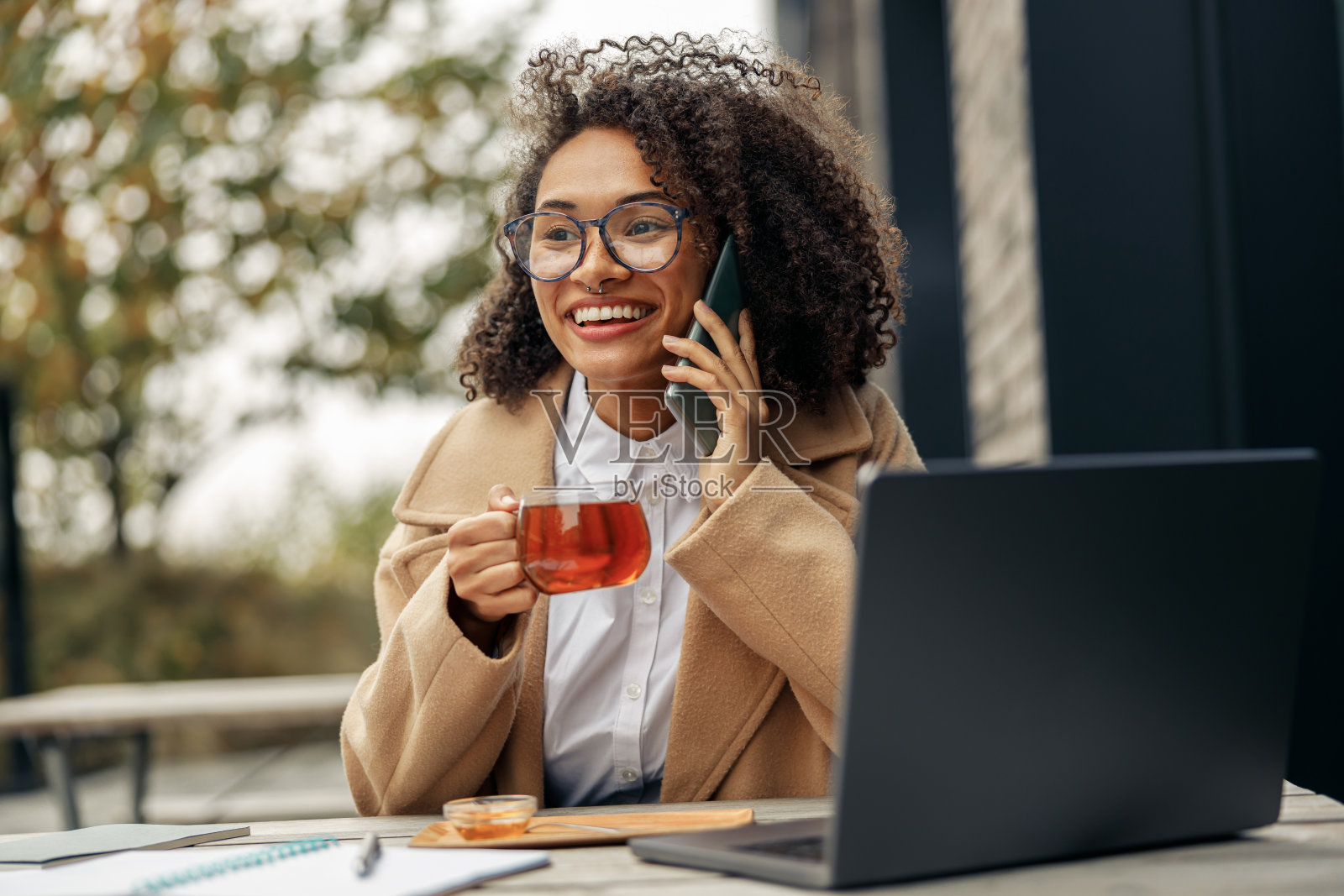 微笑的非洲裔美国女商人打电话,而工作在线坐在咖啡馆露台照片摄影图片