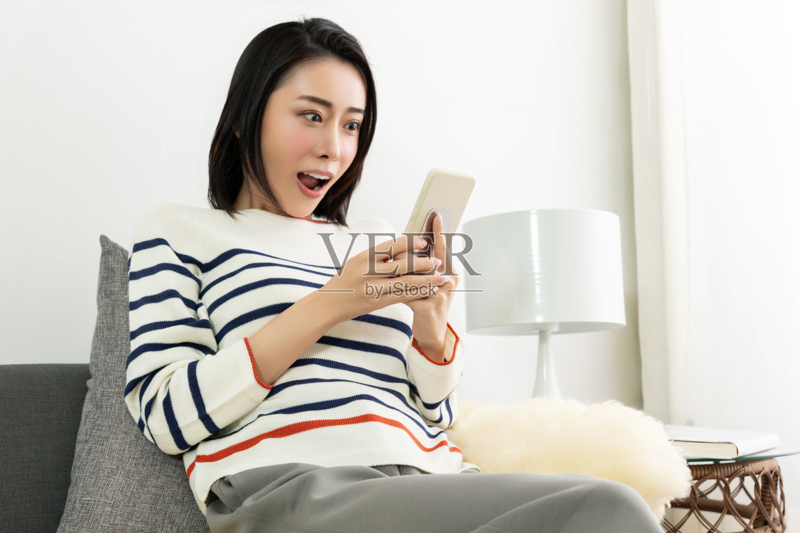 年轻漂亮的亚洲女人坐在客厅舒适的沙发上,一边用手机。照片摄影图片