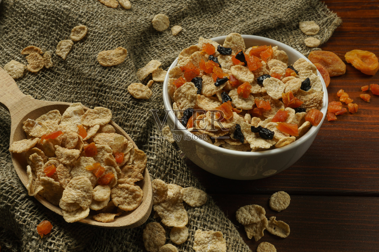 玉米片,杏干和梅干,放在一个白色的碗和一个木勺里。照片摄影图片