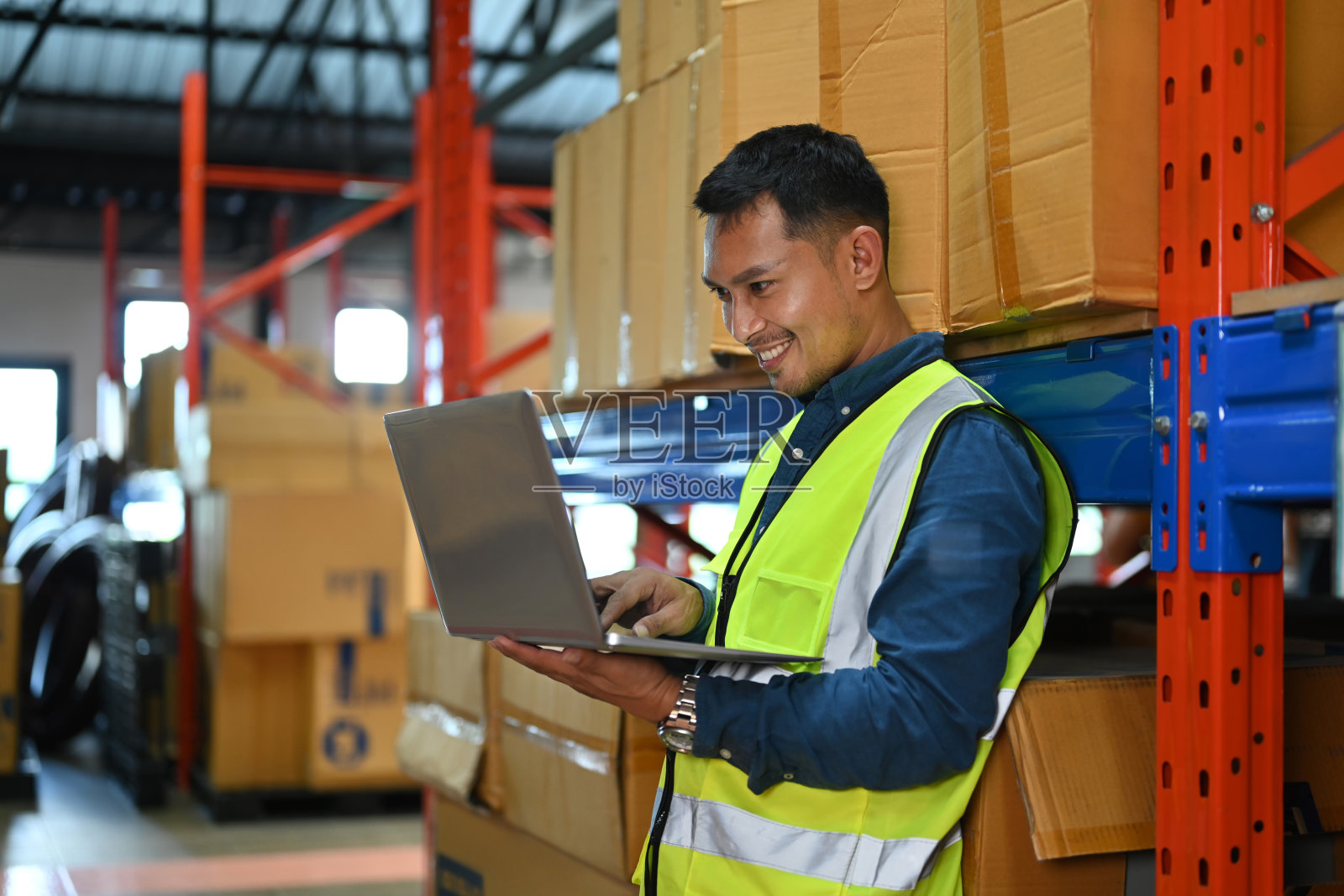 微笑的男性仓库工人使用笔记本电脑,检查货架上的库存箱在零售仓库。照片摄影图片