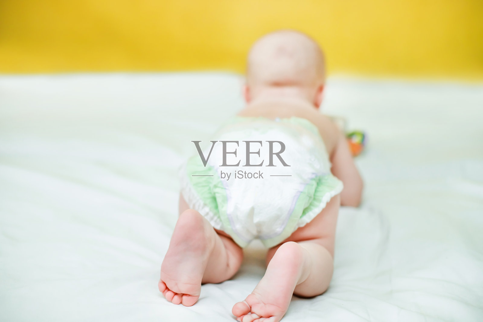 一个在水泵里爬行的孩子的后视图。婴儿卫生。防止新生儿尿流。儿童保健。可爱的宝宝躺在白色的床单上,靠在黄色的墙上照片摄影图片