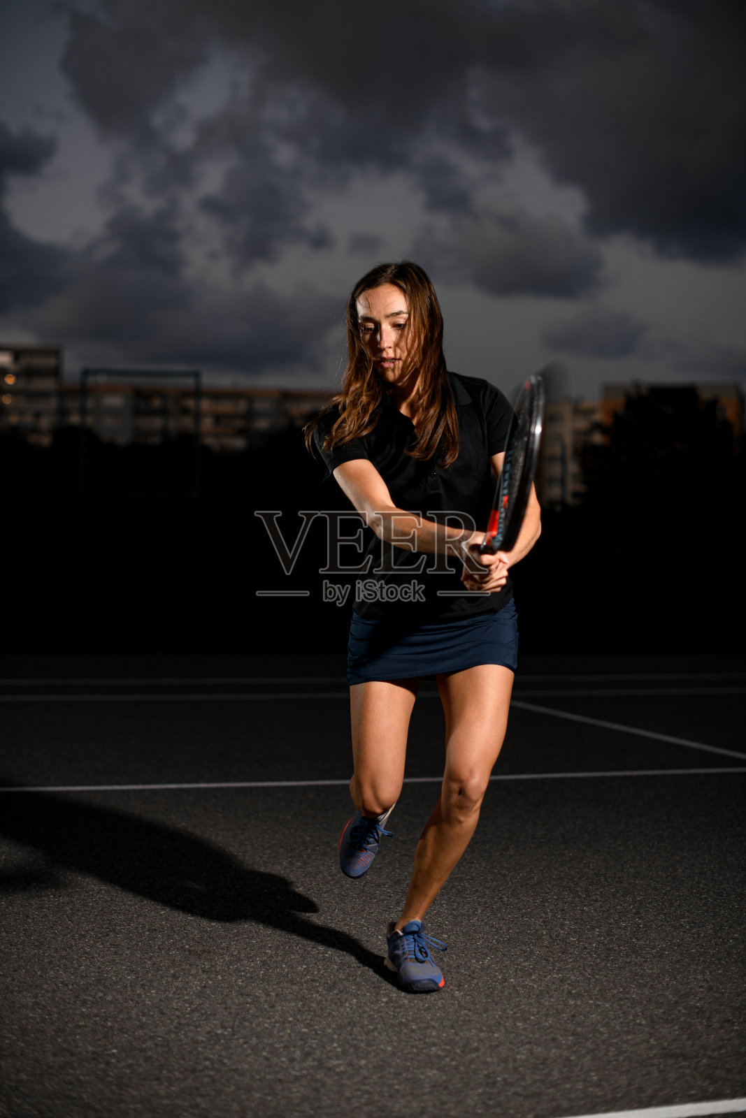 傍晚,一名女子拿着球拍在室外场地打网球。照片摄影图片