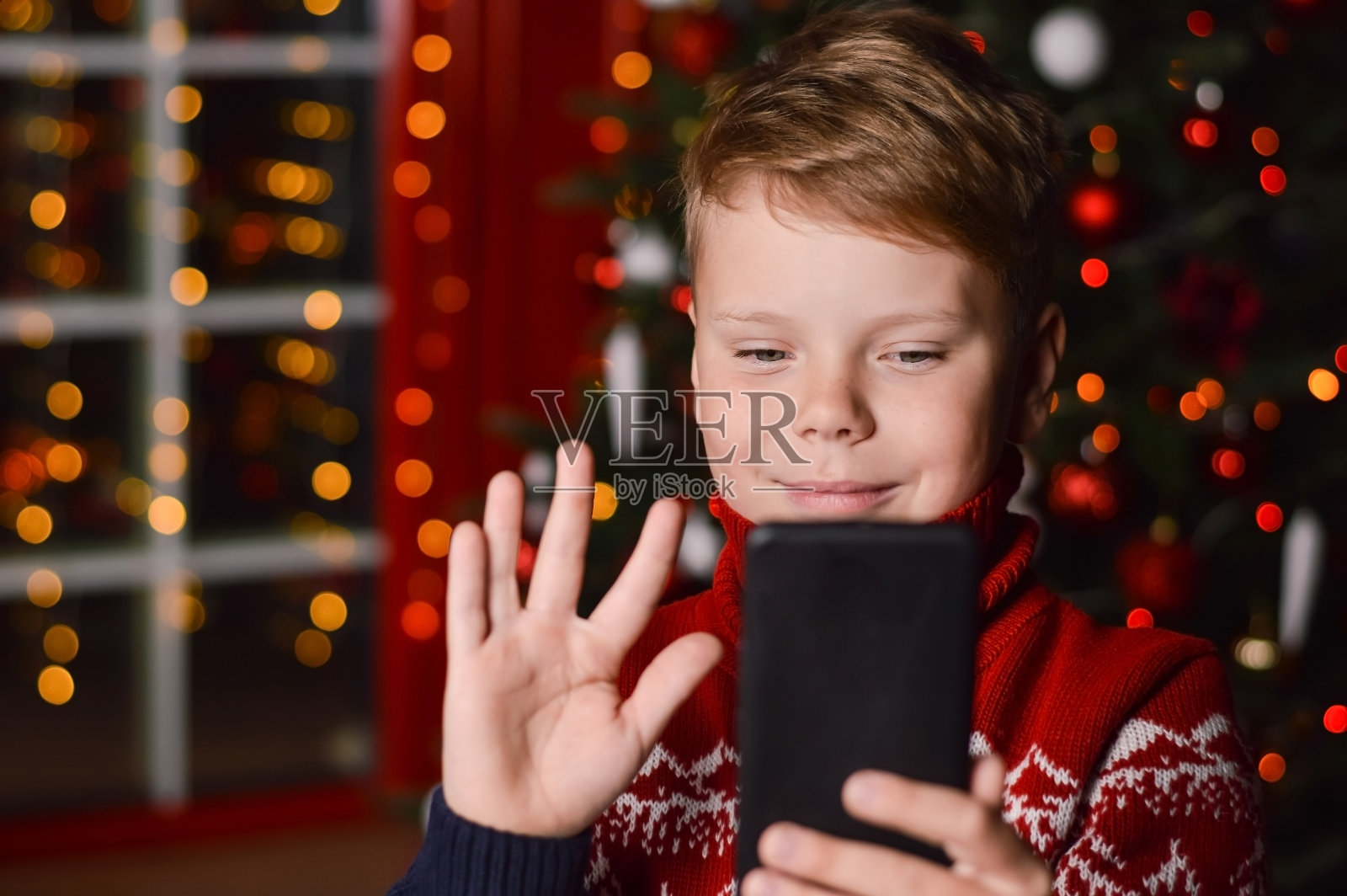 平安夜,男孩微笑着用手机拍自己的照片。圣诞树上有暗红色的灯作为背景。照片摄影图片