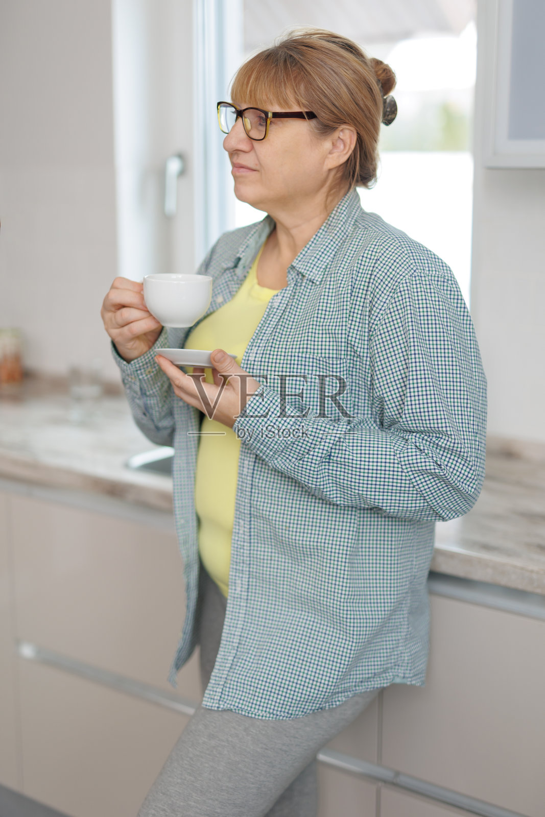 咖啡休息。冷静的高级女士在蓝色衬衫站在厨房,喝咖啡,看着空旷的空间照片摄影图片