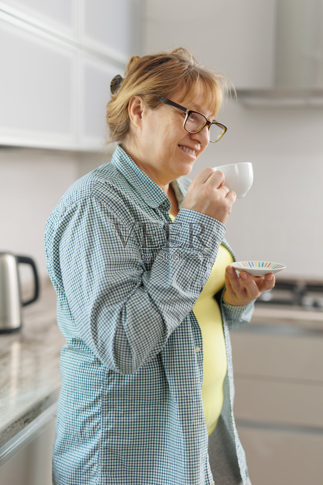 咖啡休息。冷静的高级女士在蓝色衬衫站在厨房,喝咖啡,看着空旷的空间照片摄影图片