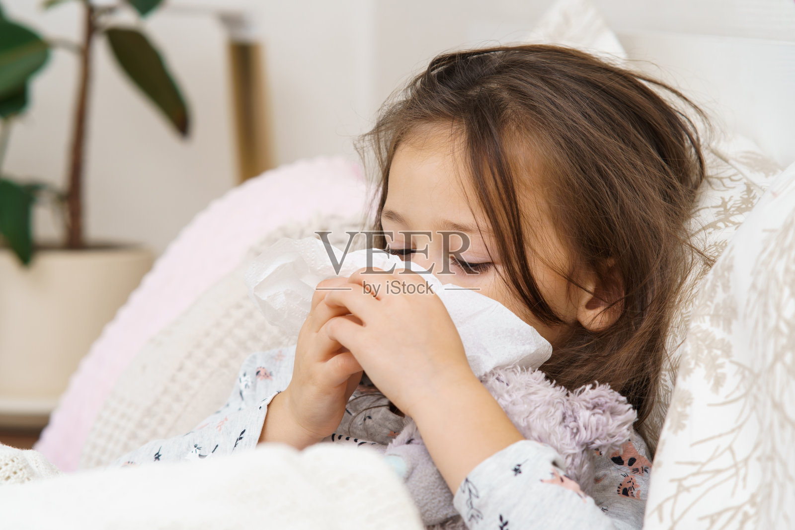 伤心沮丧的小长发女孩用纸巾捂着脸,躺着柔软的玩具,裹着毯子。过敏照片摄影图片