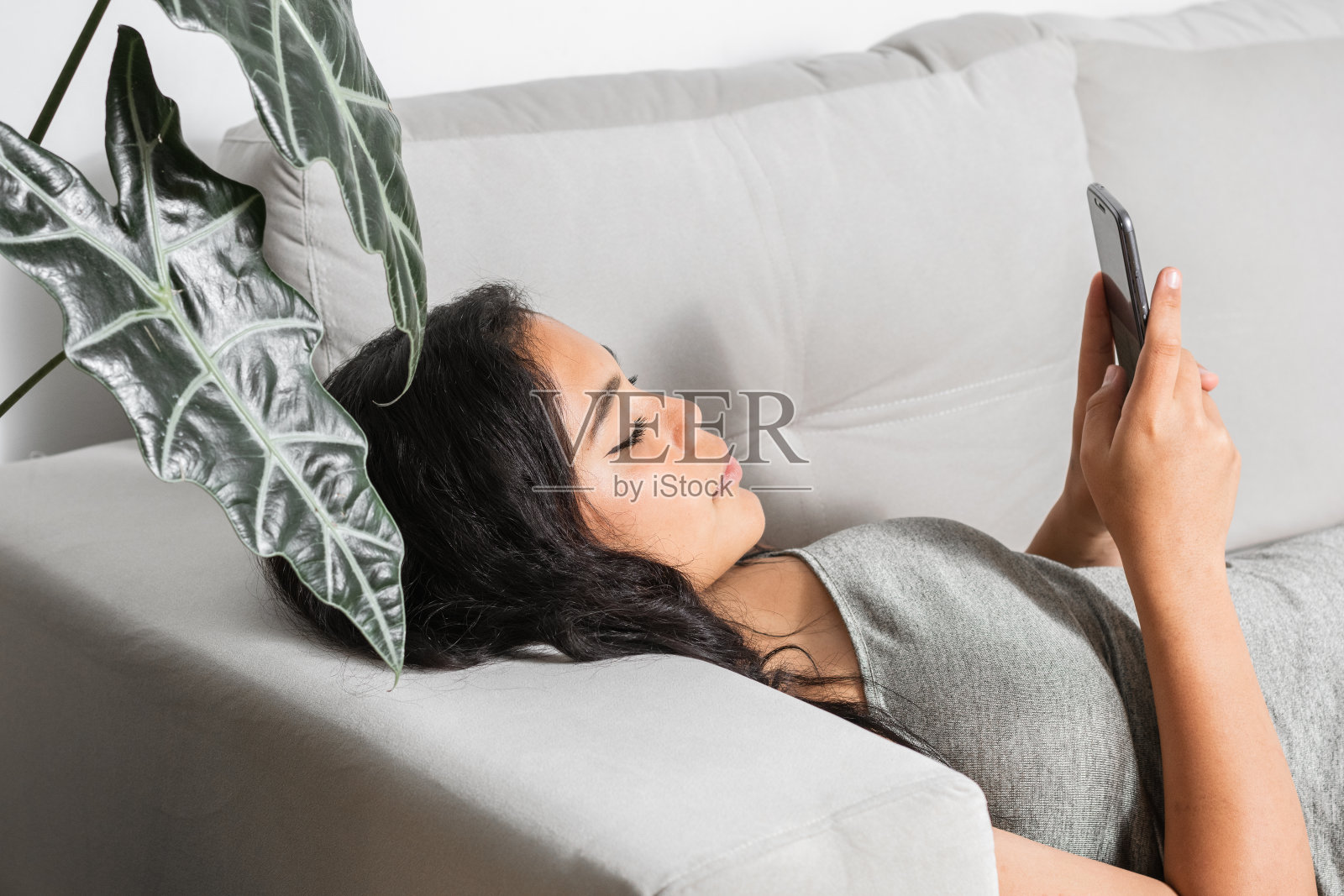 千禧一代的黑发女人,躺在沙发上看着她的手机,非常专注于她的空闲时间通过她的社交网络发送和接收消息。照片摄影图片