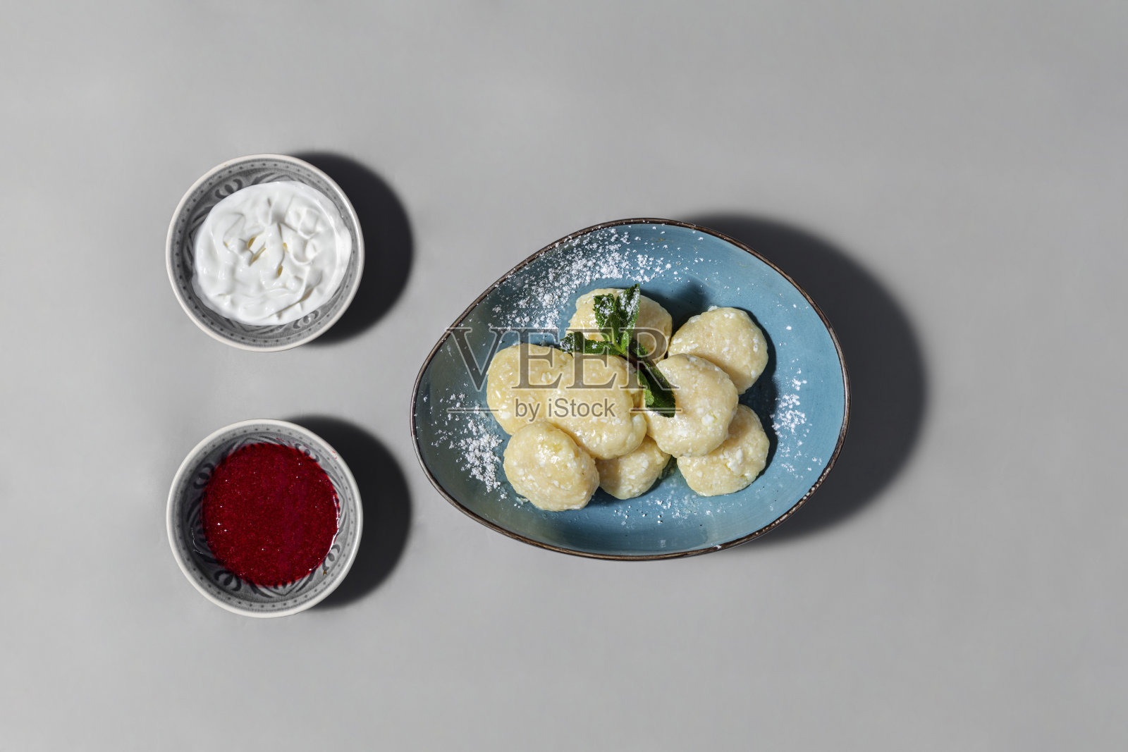 在上面撒上糖粉和薄荷叶的懒人饺子。Vareniki躺在一个蓝色的陶瓷碗里。旁边有一碗覆盆子果酱和一碗酸奶油。盘子放在灰色的纸背景上。照片摄影图片