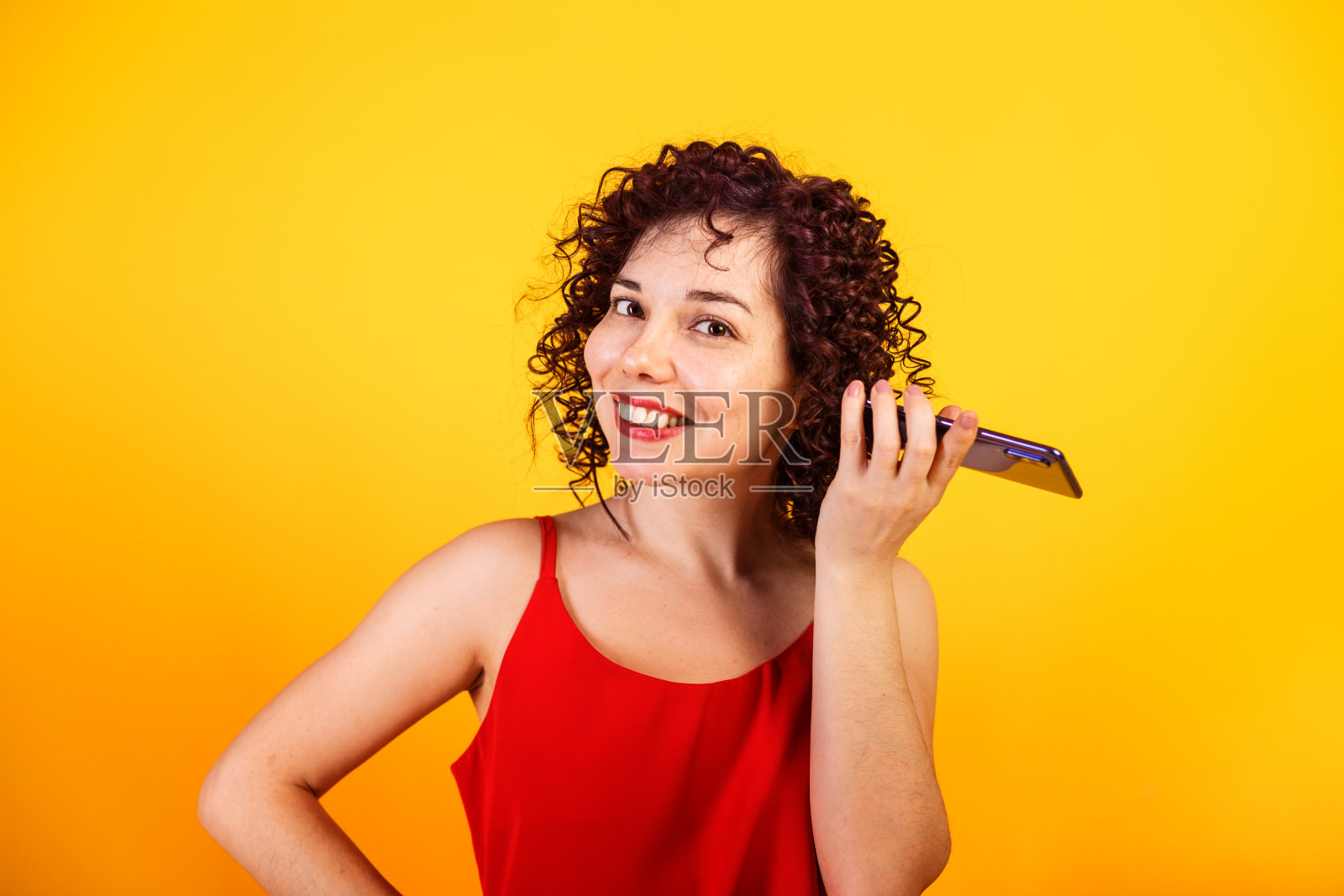 可爱的微笑卷发女孩正在打电话。一位年轻女士正在听语音信息。工作室女性肖像在明亮的黄色背景。照片摄影图片