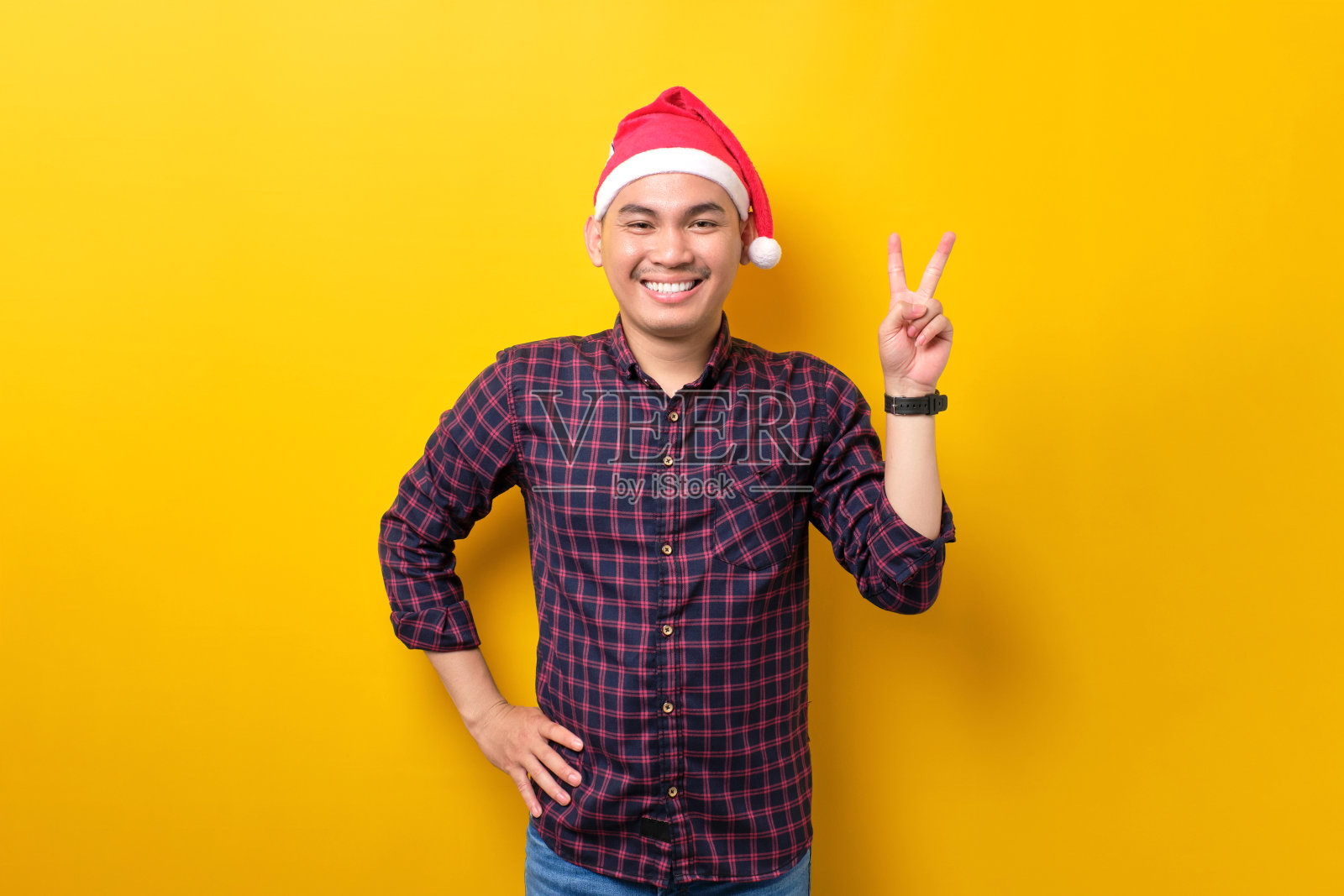 快乐的年轻亚洲男子戴着圣诞老人的帽子,做出胜利的手势,看着黄色工作室背景上的相机,新年快乐2023庆祝节日快乐概念照片摄影图片