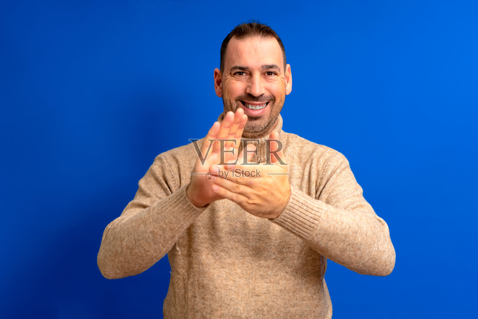 大胡子西班牙男子穿着棕色高领毛衣在孤立的蓝色工作室背景拍手高兴和开朗,微笑自豪与双手在一起。照片摄影图片