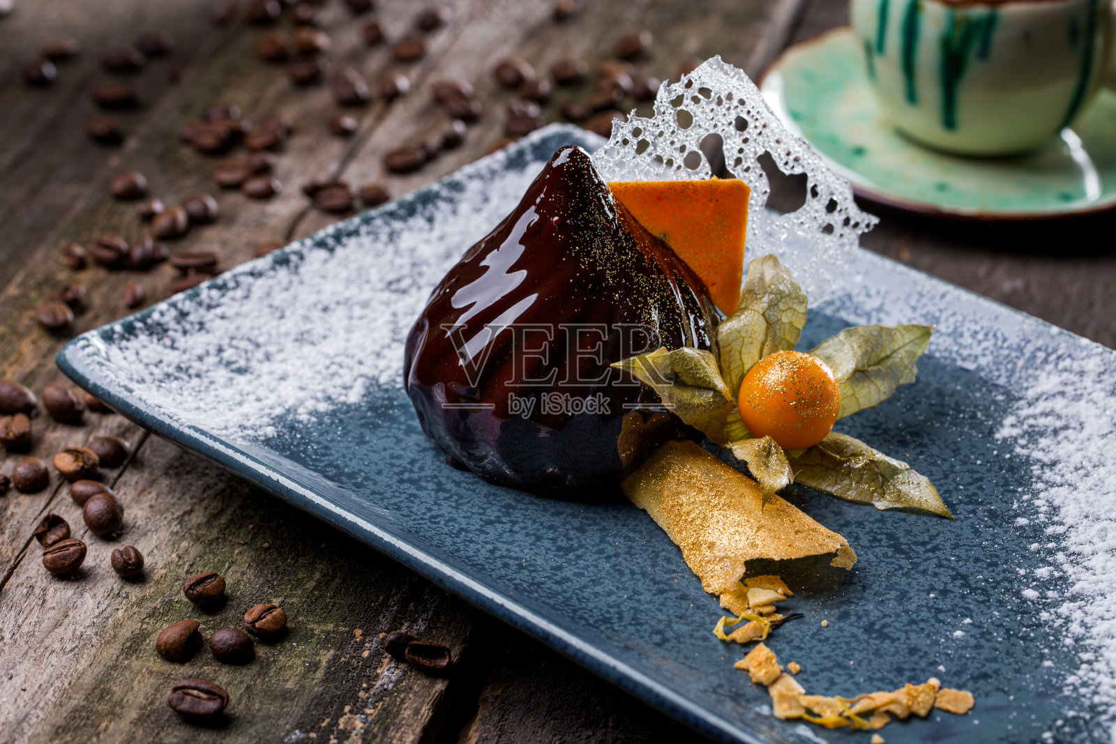 巧克力釉甜点,有酸浆,透明的薄糖釉和酥皮,糖粉,金色的糖粉洒在蓝色的盘子上,木桌上散落着咖啡豆。水平方向照片摄影图片