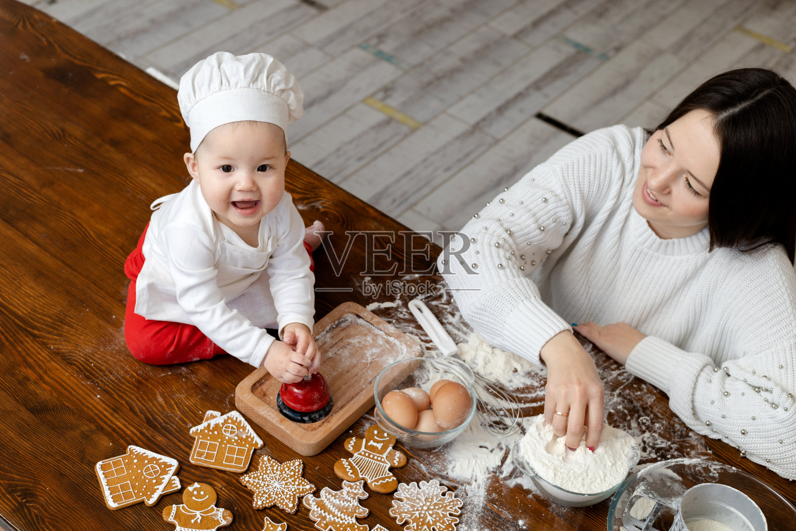 一个戴着厨师帽和围裙的亚洲婴儿和年轻的妈妈在家里厨房的桌子上做圣诞姜饼,开心地笑着照片摄影图片