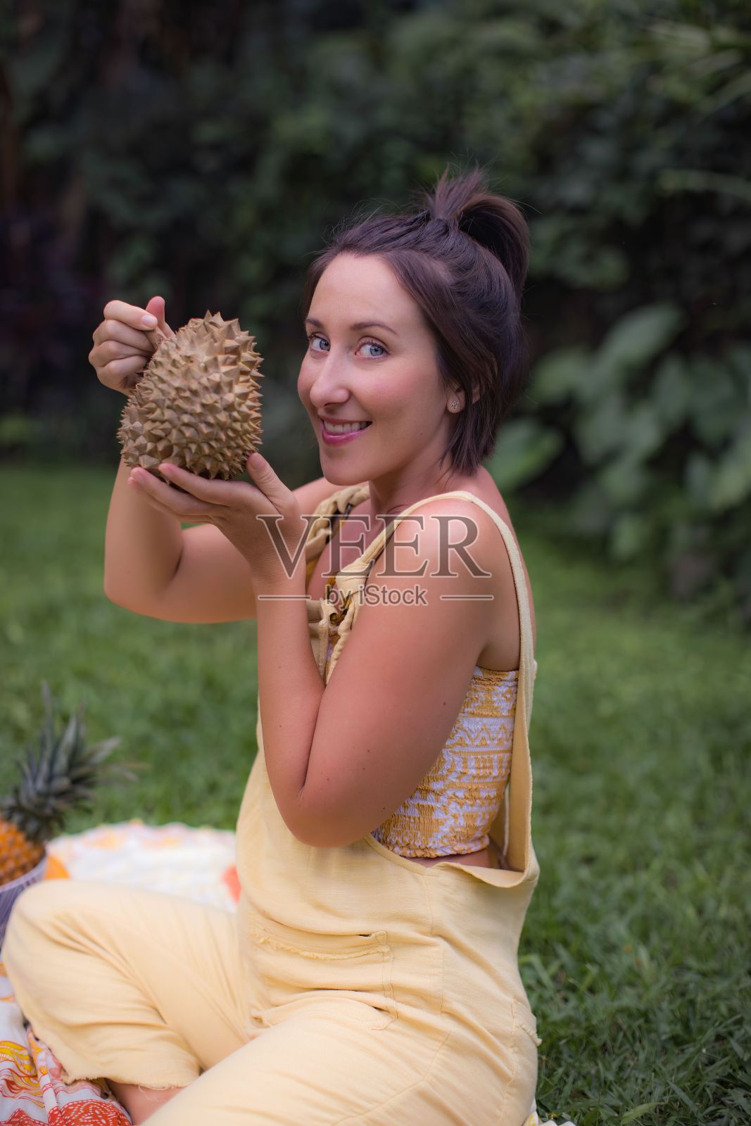 垂直特写,一位美丽的年轻女子坐在草坪上,手里拿着榴莲。夏季野餐。照片摄影图片
